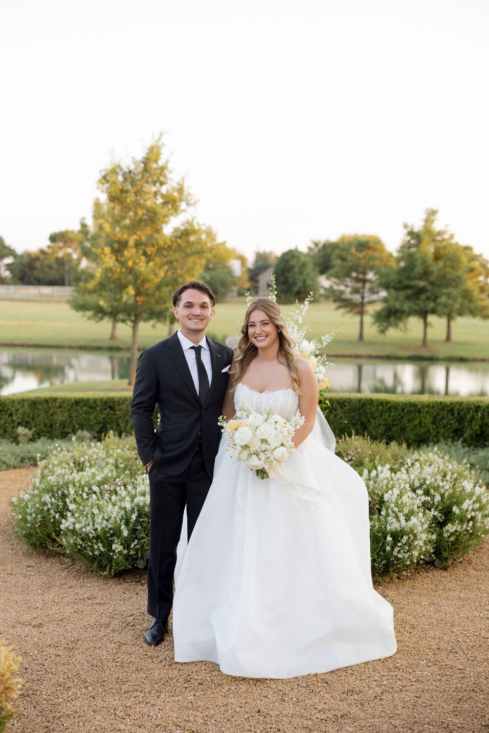 A bride and groom pose for portraits in a manicured garden landscape with white flowers and autumn trees.