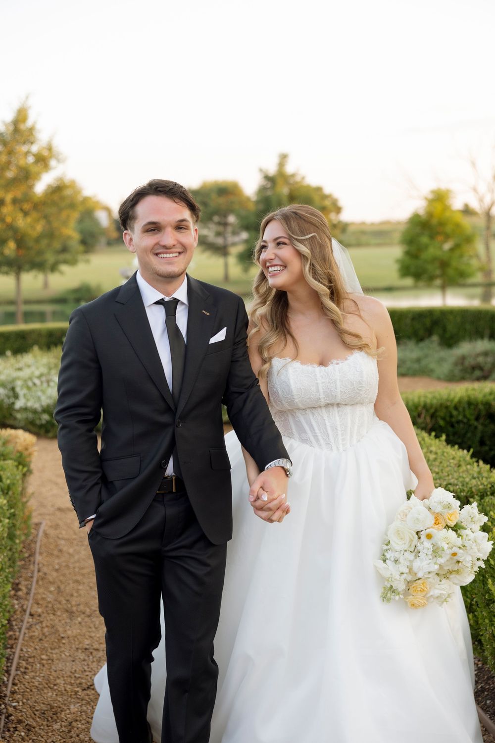 Joyful wedding couple walks hand in hand along garden path at golden hour.