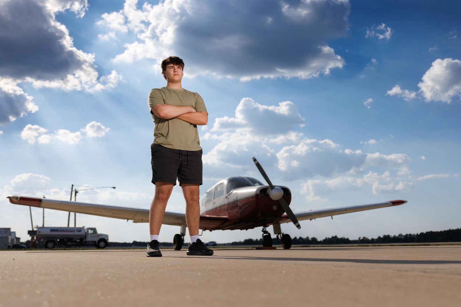A person stands confidently in front of a small private aircraft on a sunny day with dramatic clouds in the sky.