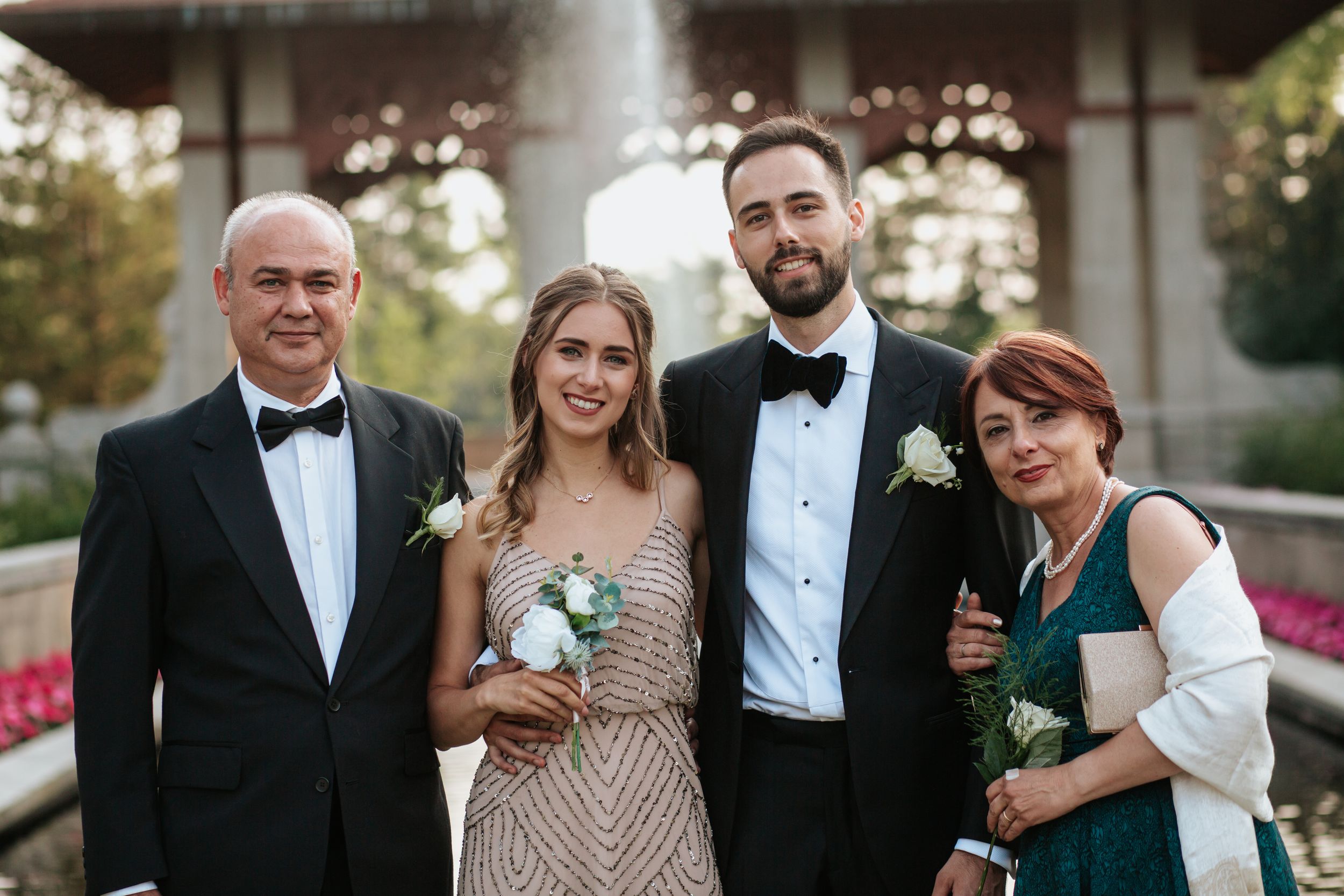 Formal wedding party members in black tuxedos and elegant dresses pose together during an outdoor evening celebration.