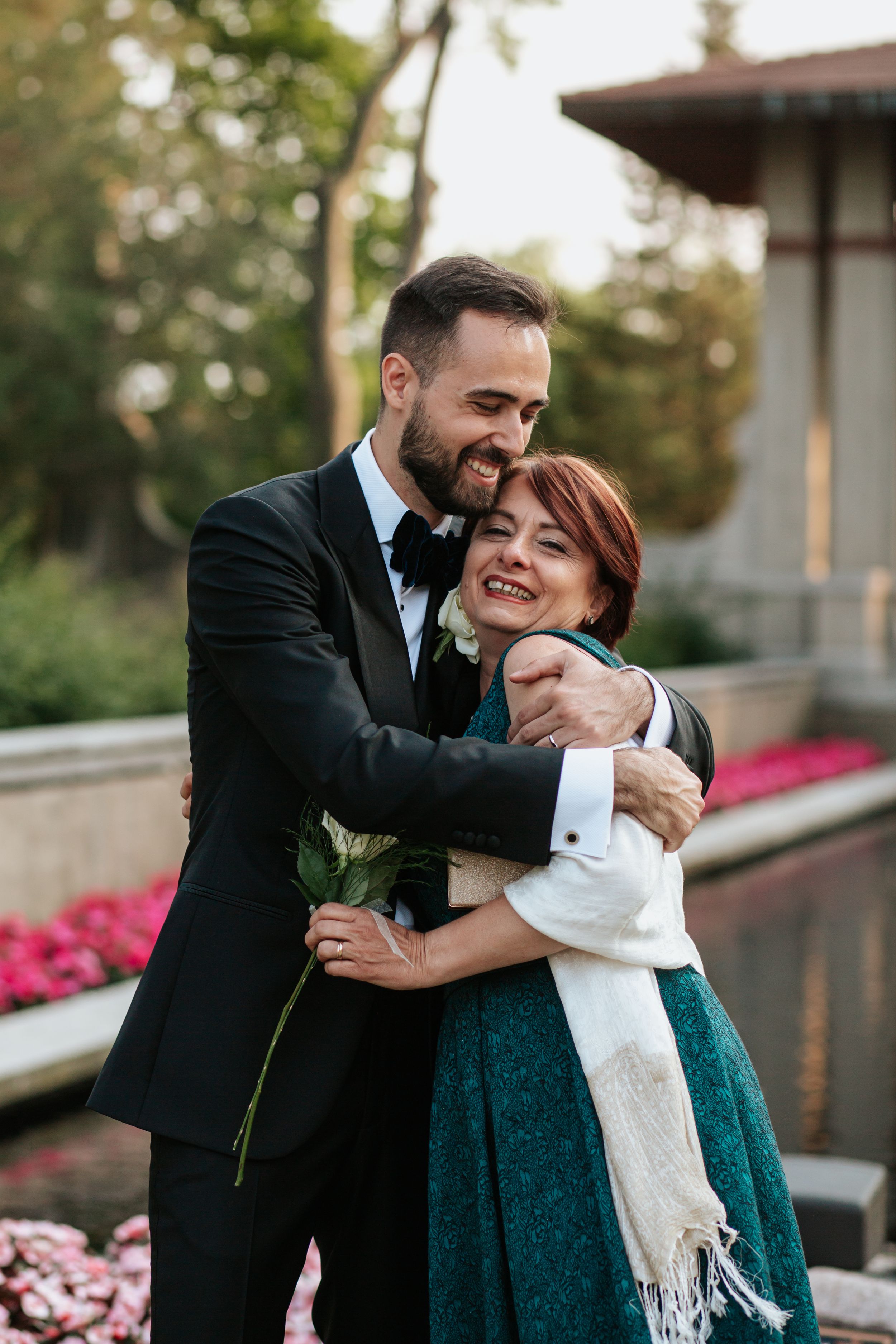 A couple shares a joyful embrace outdoors with the person in teal dress and white scarf being hugged warmly.