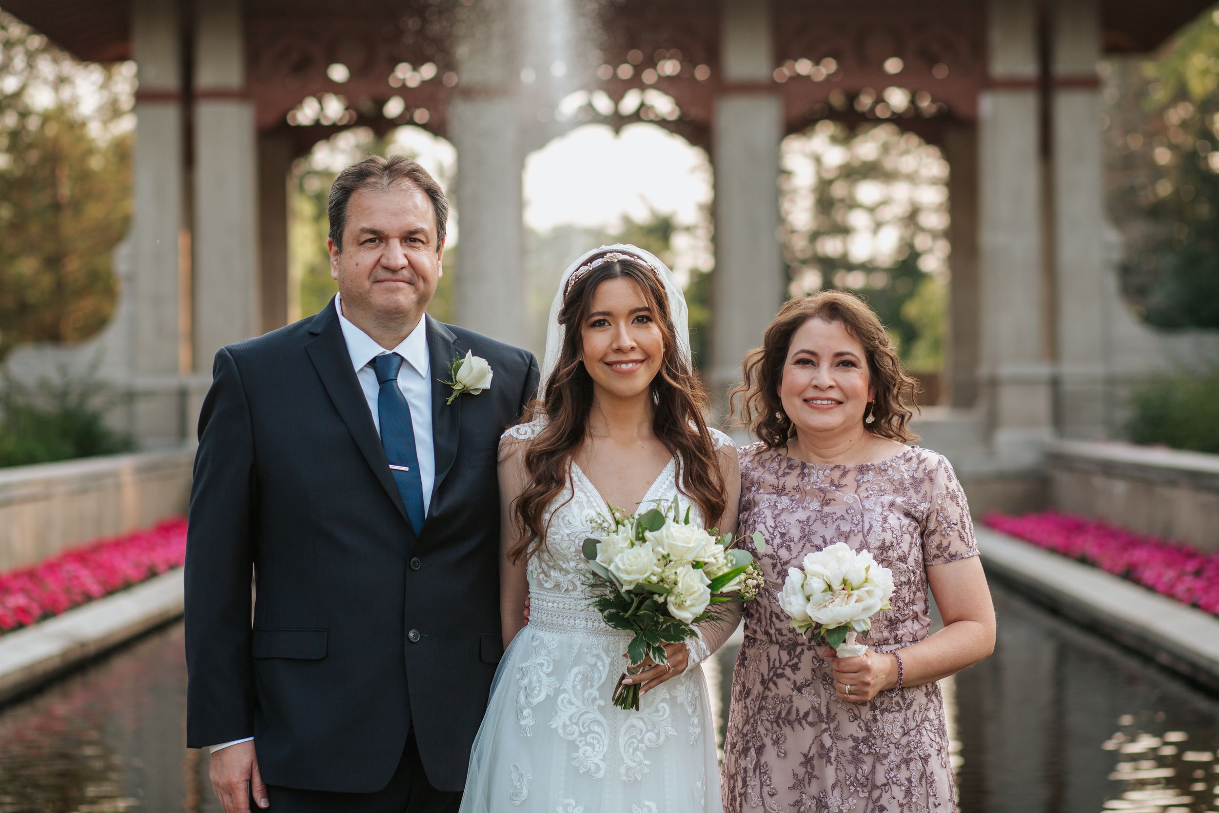 A wedding party poses together in formal attire on a sunny day at an elegant outdoor venue with blooming flowers.