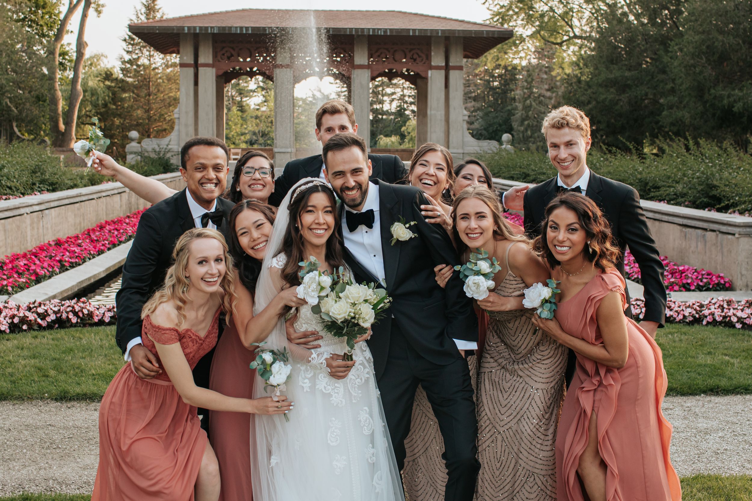 Wedding party members pose together outdoors in formal attire with coral and blue dresses and dark suits.