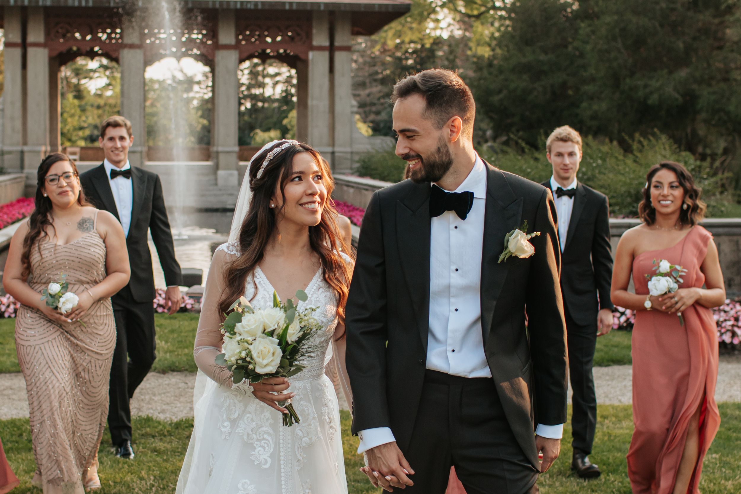 Wedding party laughs and celebrates together outdoors during sunset with elegant attire and floral bouquets.