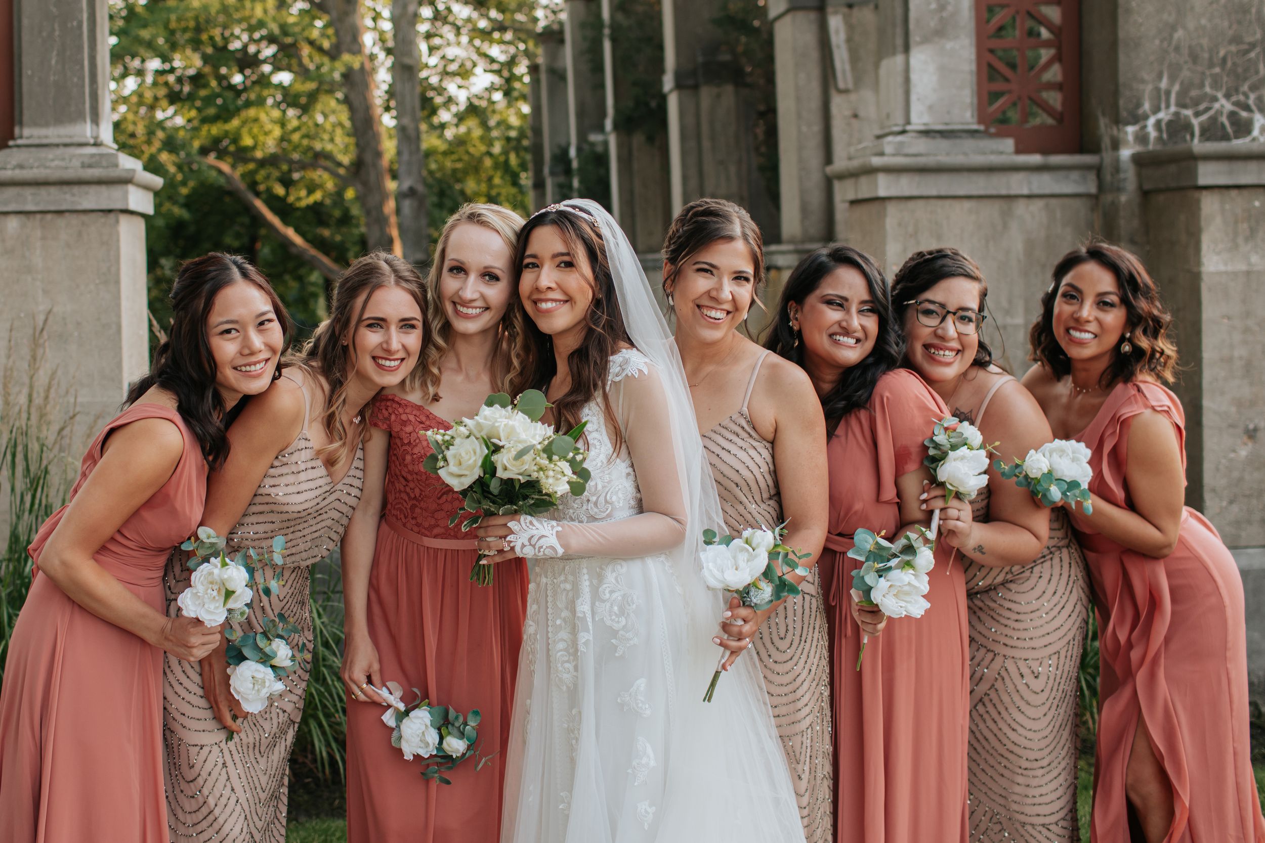 A wedding party poses together in coral bridesmaid dresses and formal attire outside a stone building.