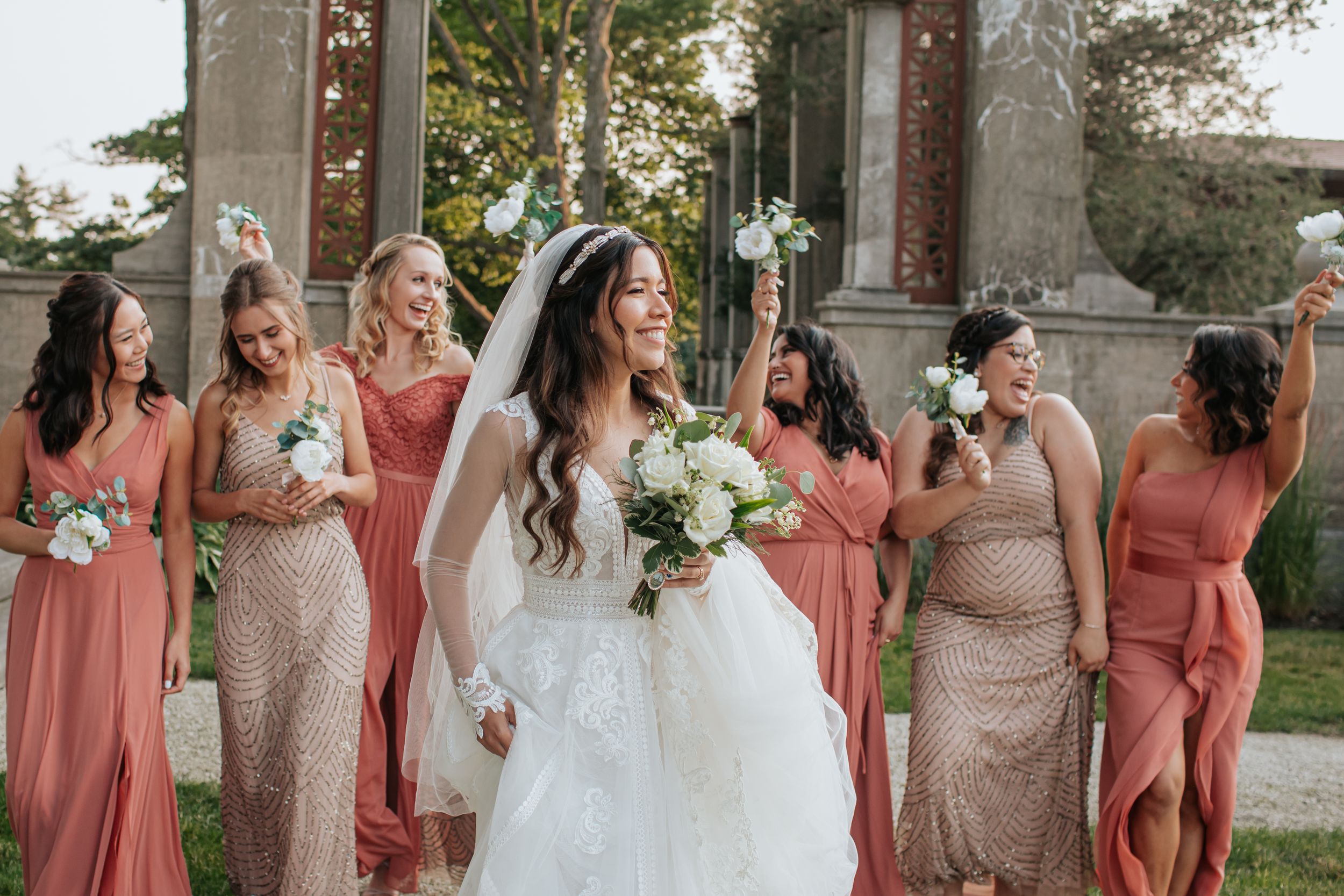 A wedding party poses in coordinating coral and champagne dresses in an outdoor garden setting.