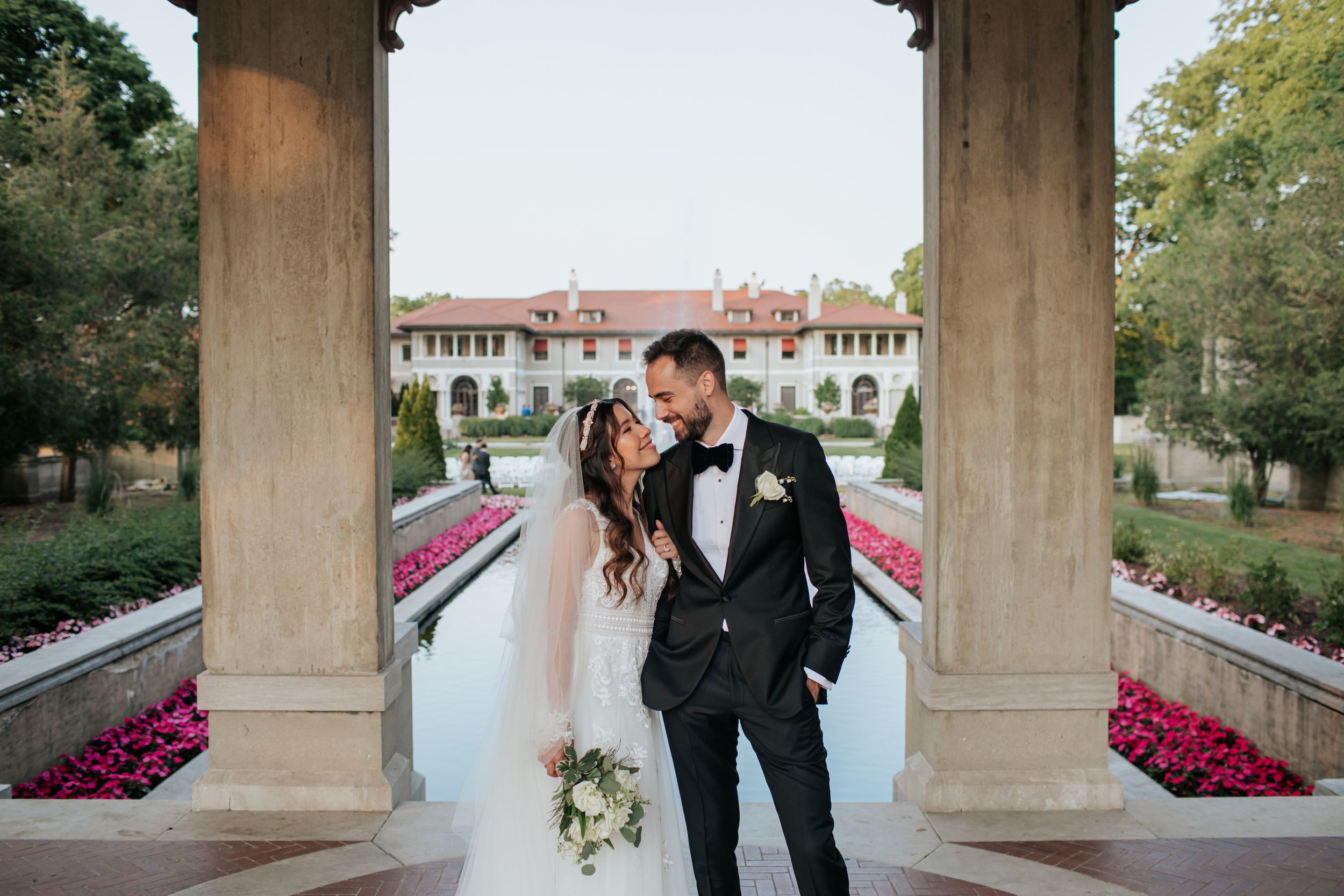 A couple walks together through stone archways with pink flowers and water features at an elegant garden venue.