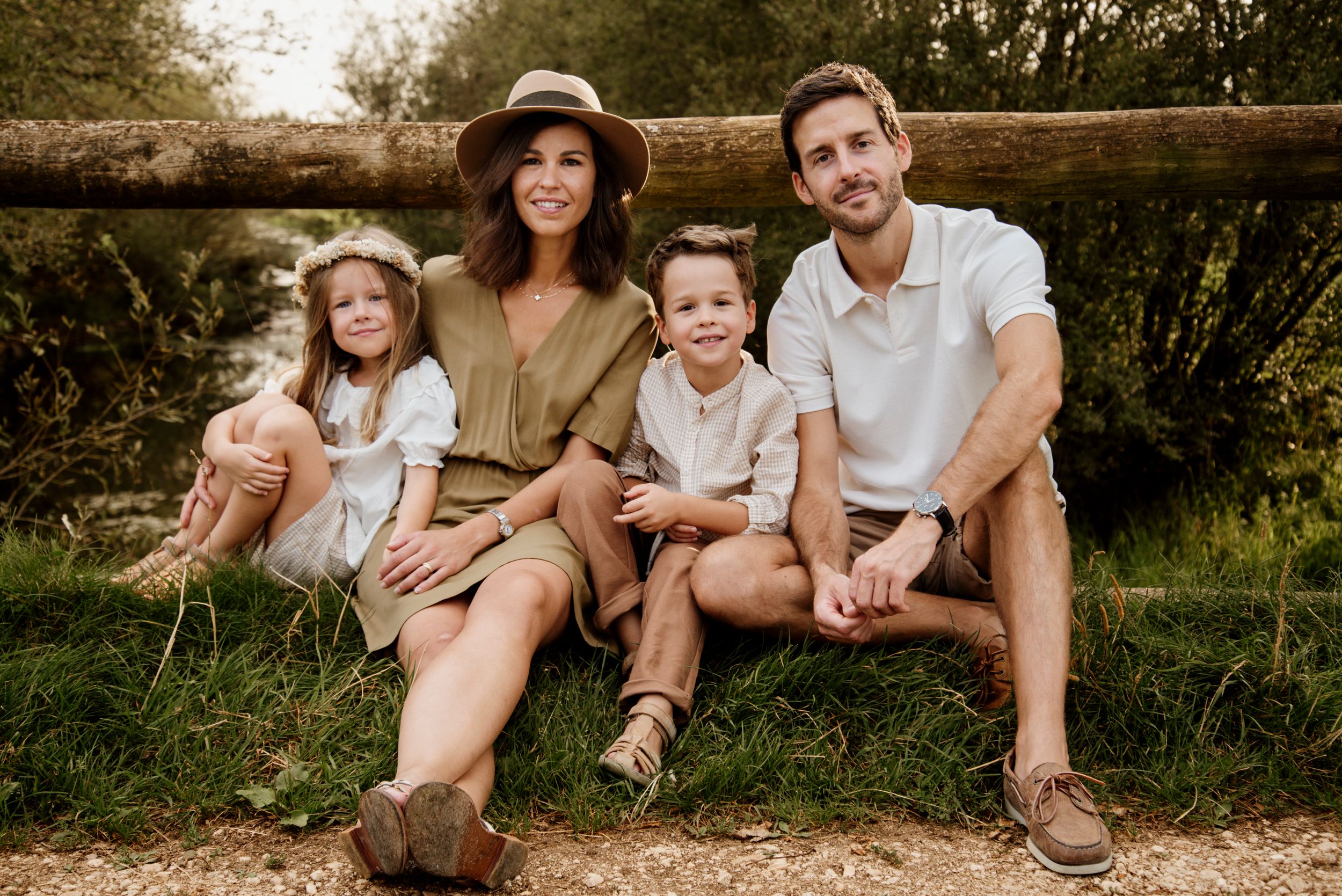 A smiling family of four sits together on a wooden fence in a sunny outdoor setting.