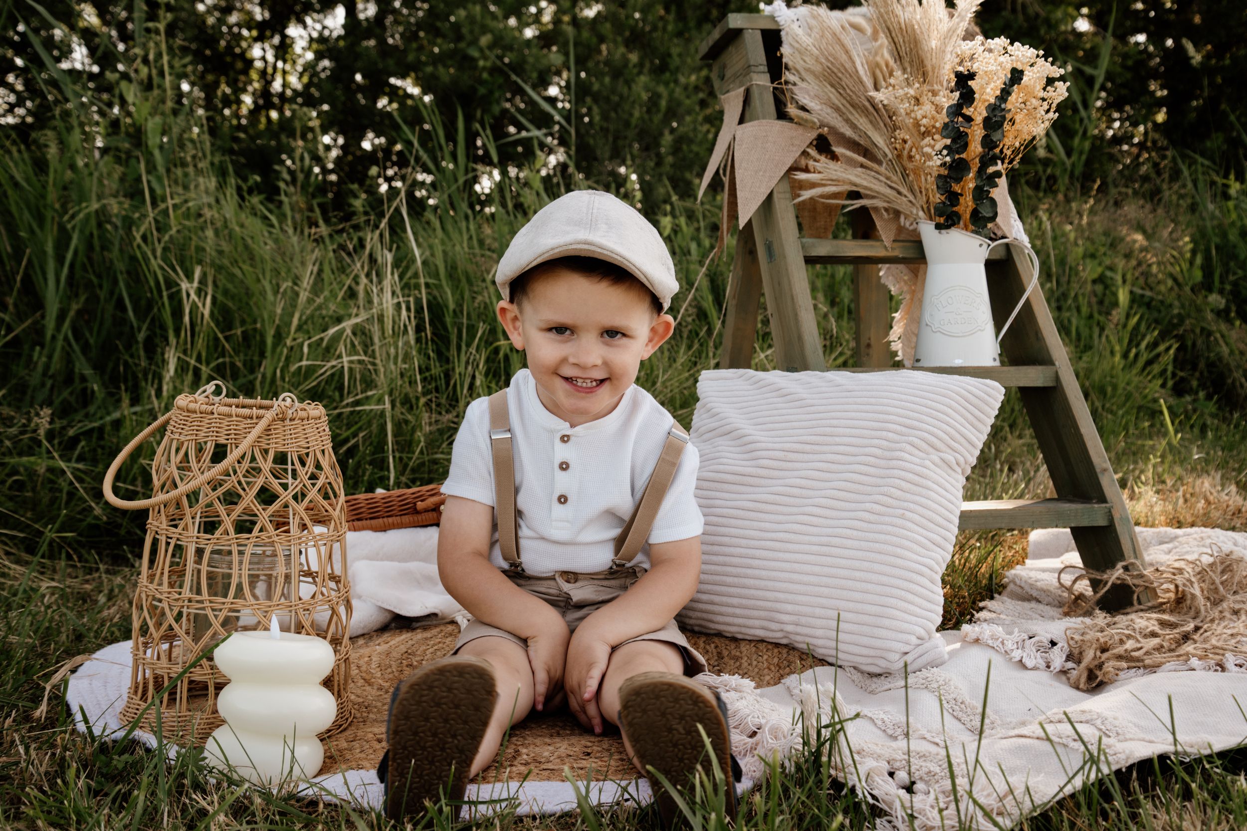 Adorable toddler boy in white cap and suspenders sitting outdoors among boho picnic decor in a meadow.