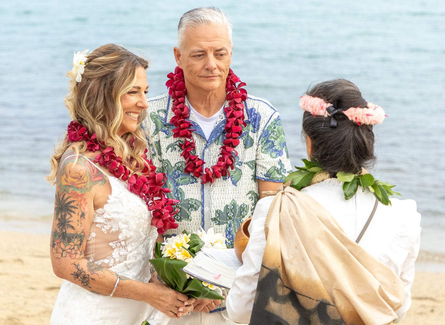 Newlywed couple shares a romantic kiss on a tropical beach with ocean waves in the background.