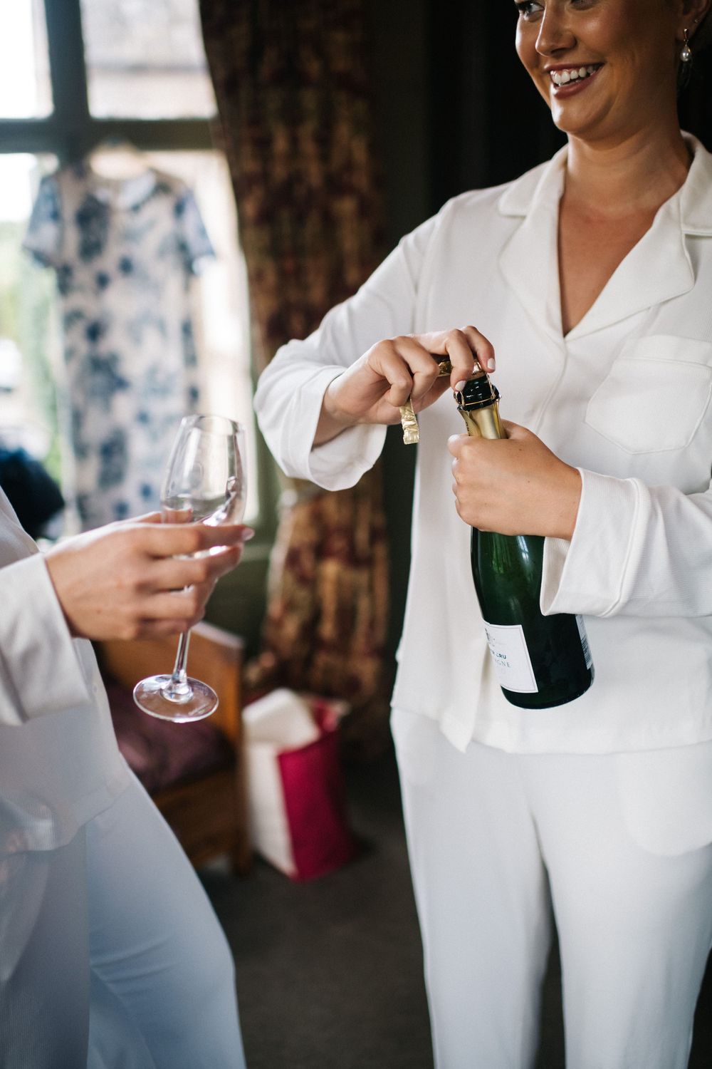 Someone in a white suit opens a bottle of champagne while another person holds a wine glass.