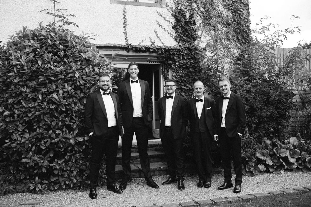 Groomsmen in black tuxedos pose together in formal wedding party portrait.