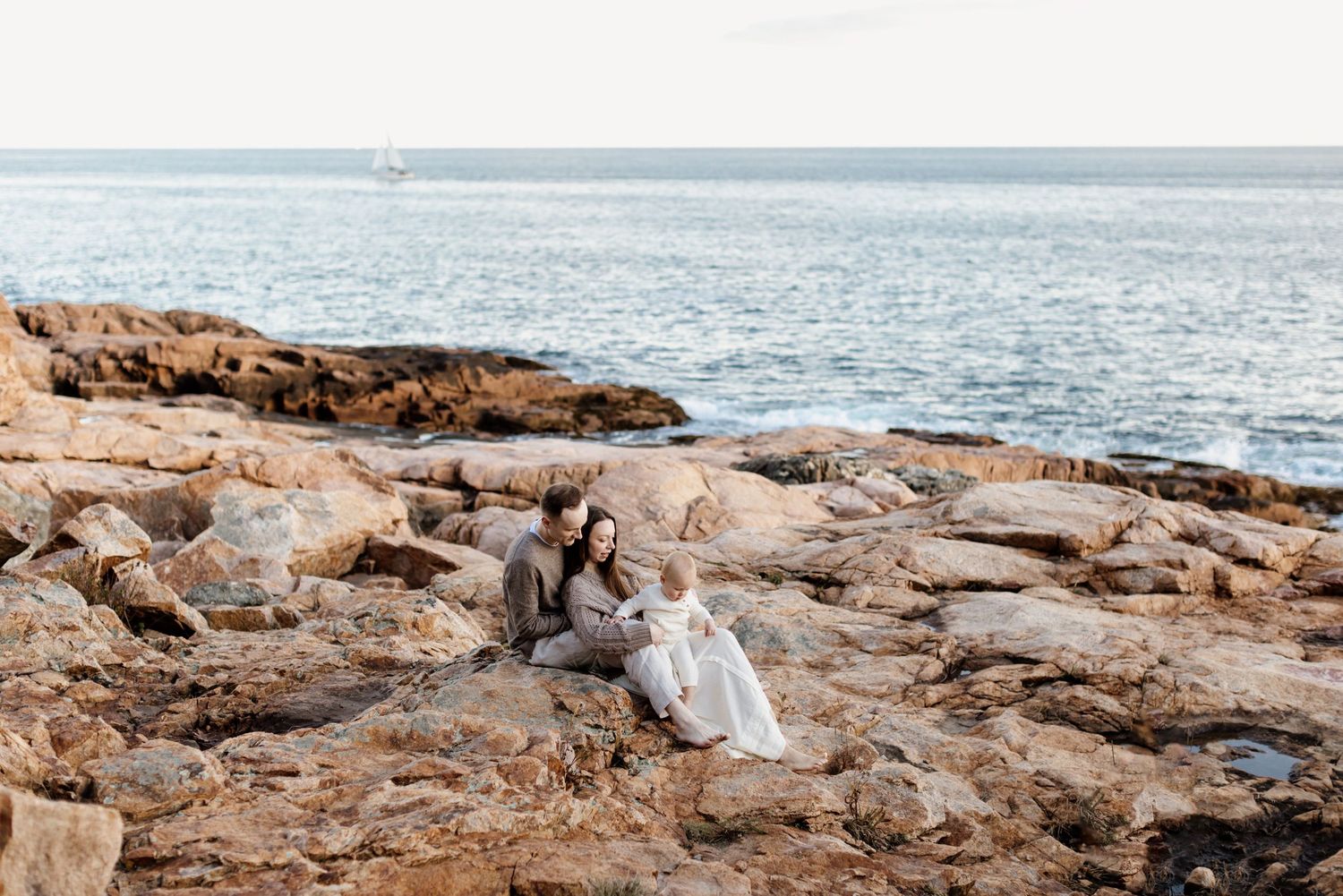 A husband and wife with a baby lounge on a golden coastal rocks with the blue ocean in the background for family photos in Gloucester, MA.