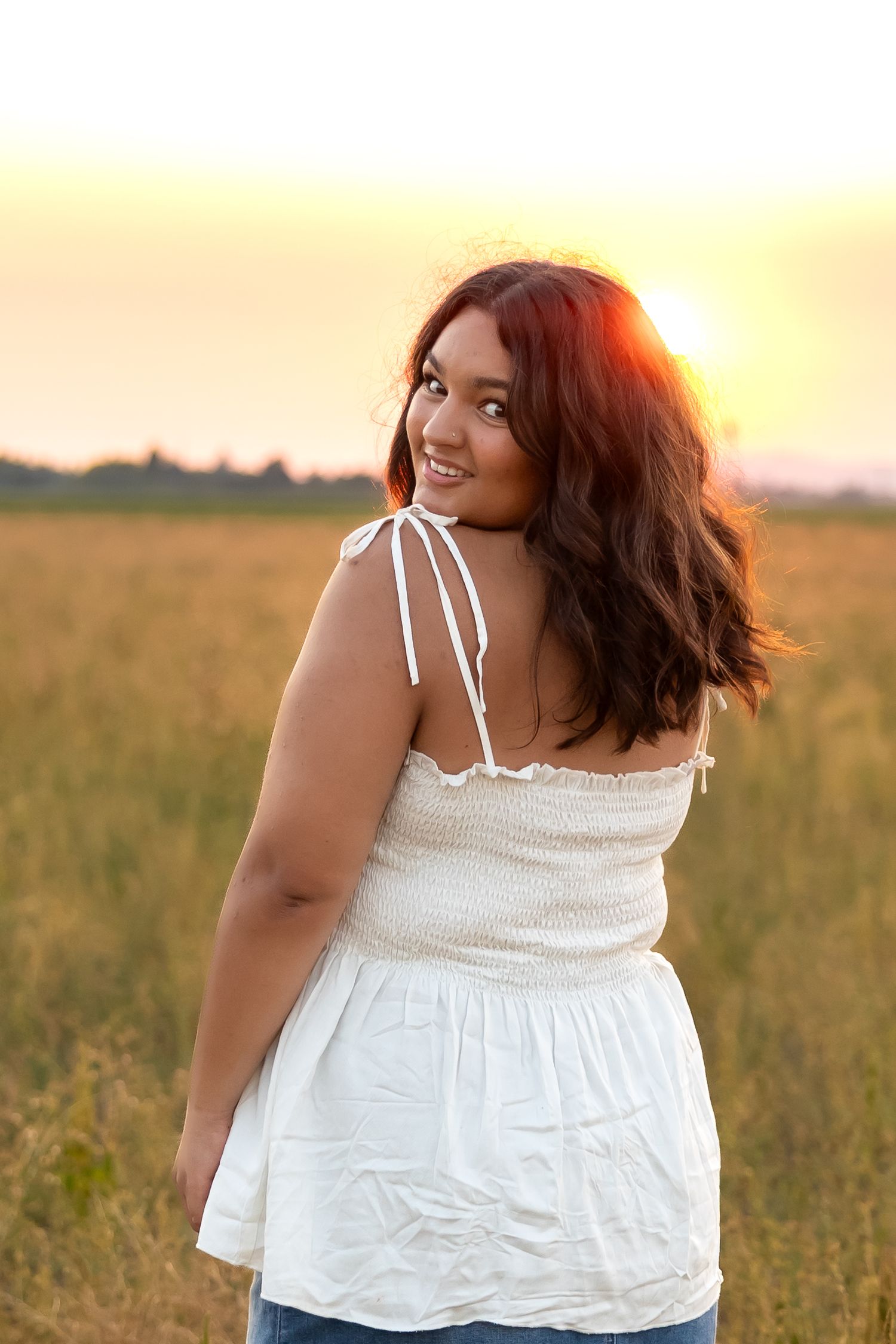 A person wearing a white summer top poses in a golden field at sunset.