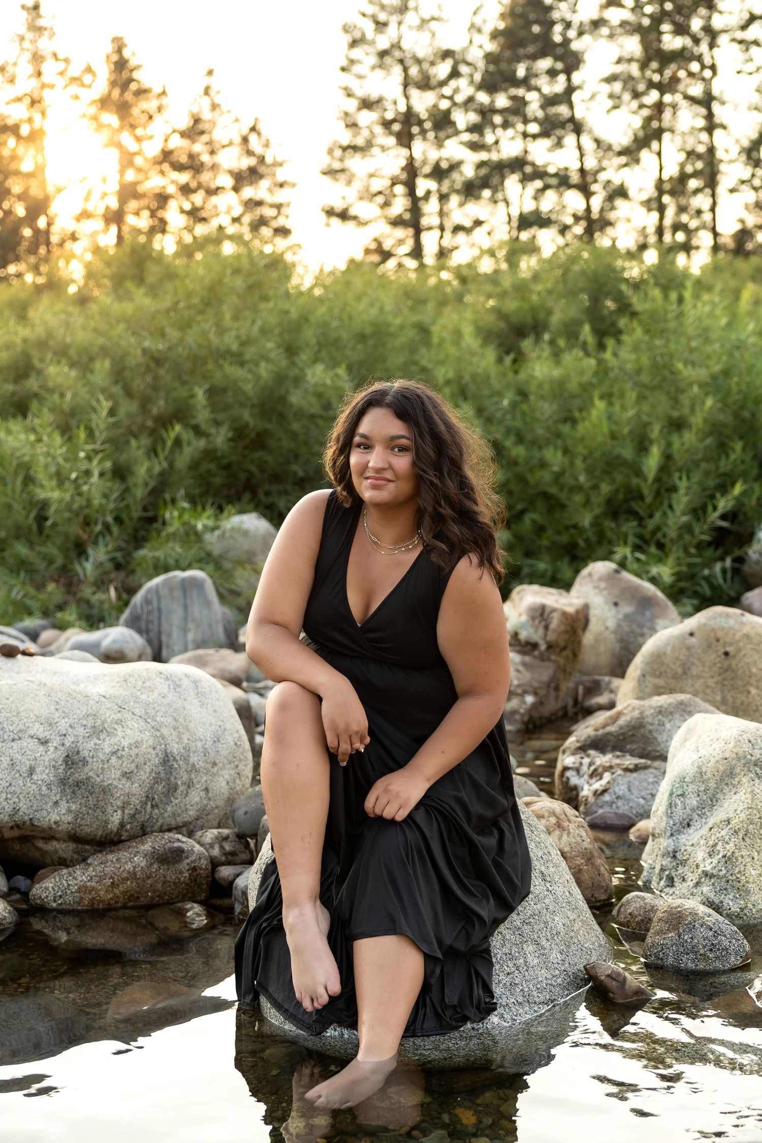 A person in a black dress poses on rocks near water during golden hour sunset.