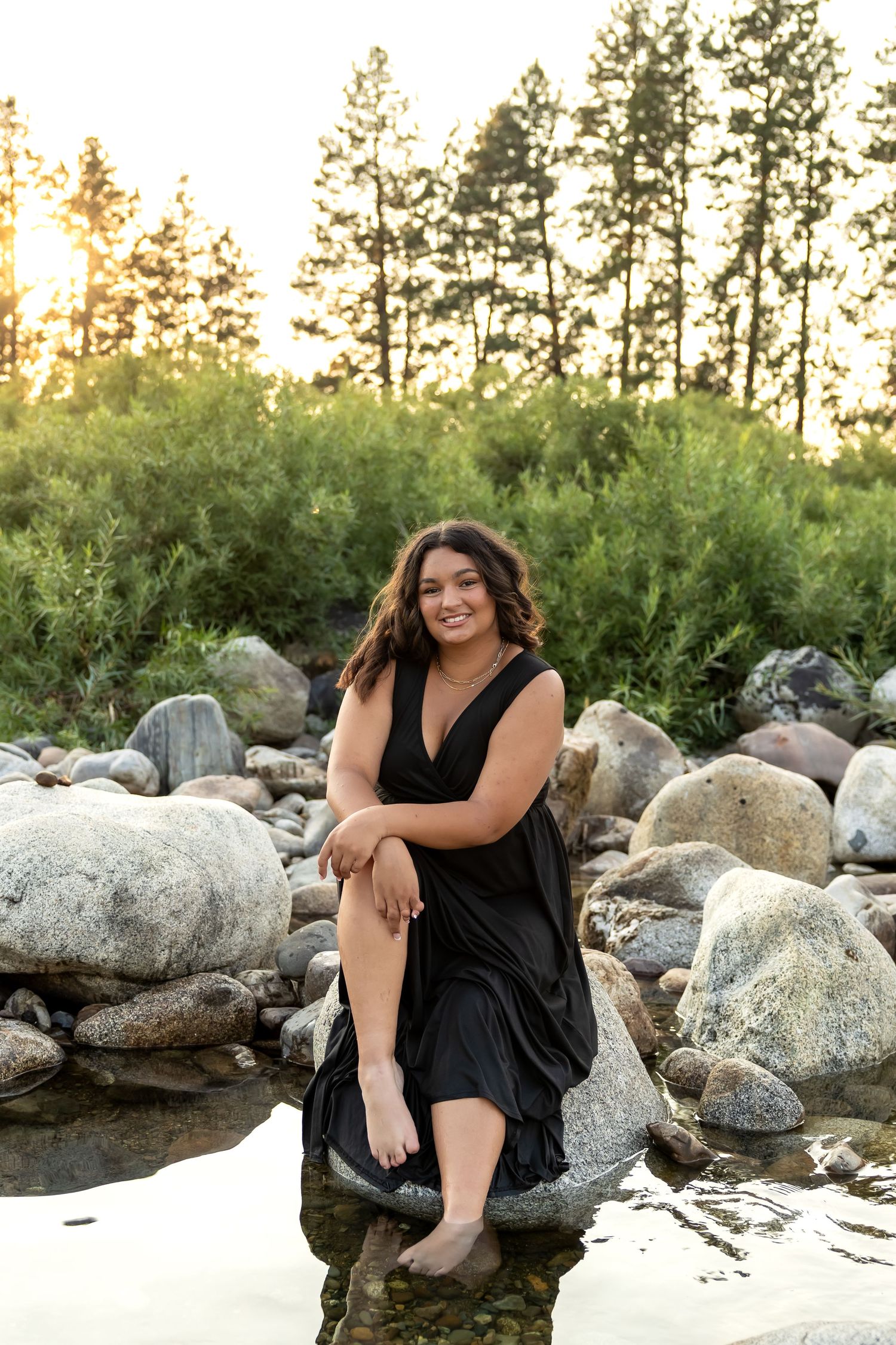 A black dress photoshoot by a rocky riverbank with tall pine trees in the background at sunset.