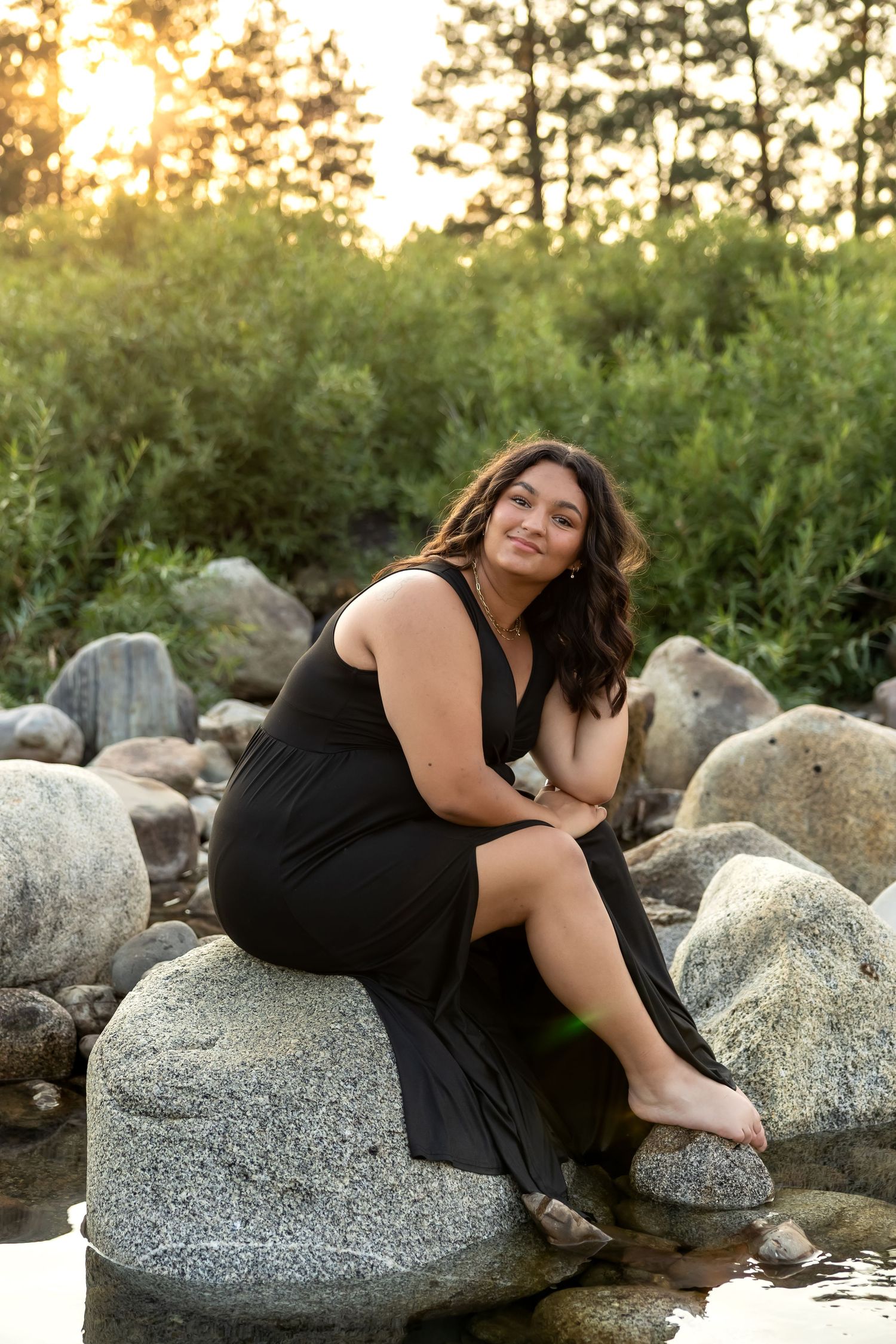 A person in a black dress poses gracefully on granite rocks near a lake at sunset.