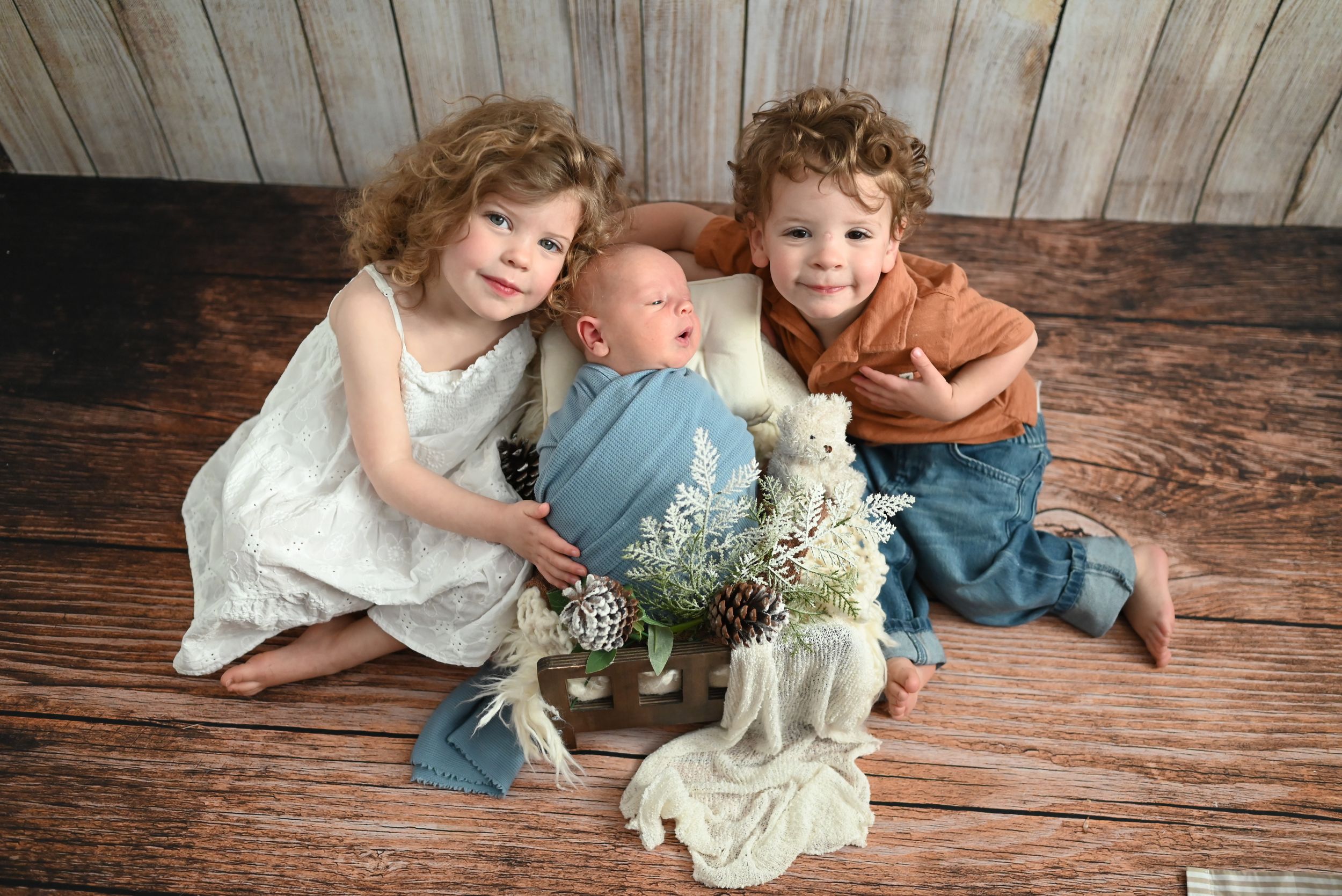Children in coordinated blue and white outfits pose together on rustic wooden flooring with floral arrangements.