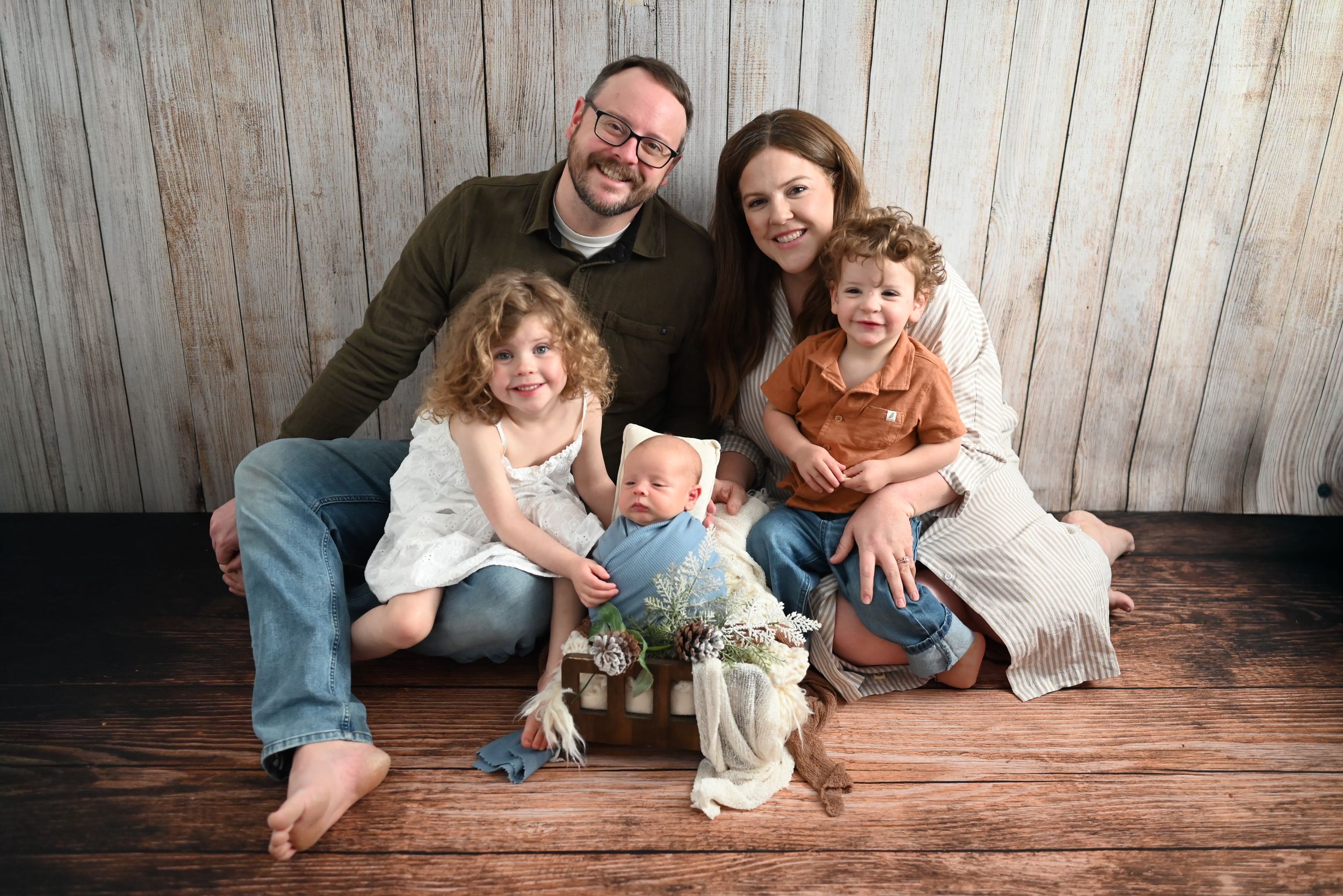 A family poses together on a wooden floor against a rustic wall with a small plant centerpiece.