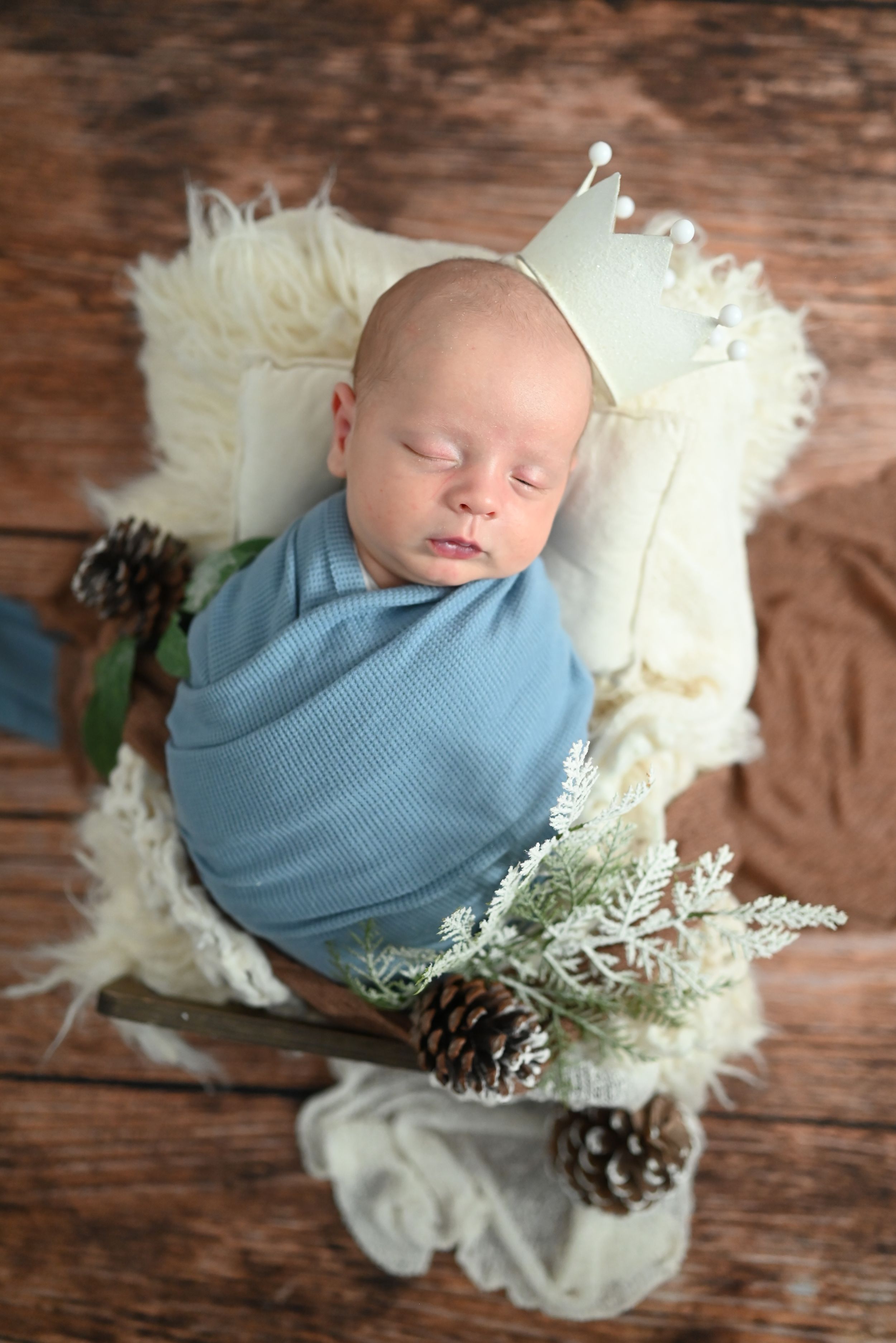 Sleeping newborn wrapped in soft blue swaddle rests on white fur with pinecones and greenery on wooden backdrop.