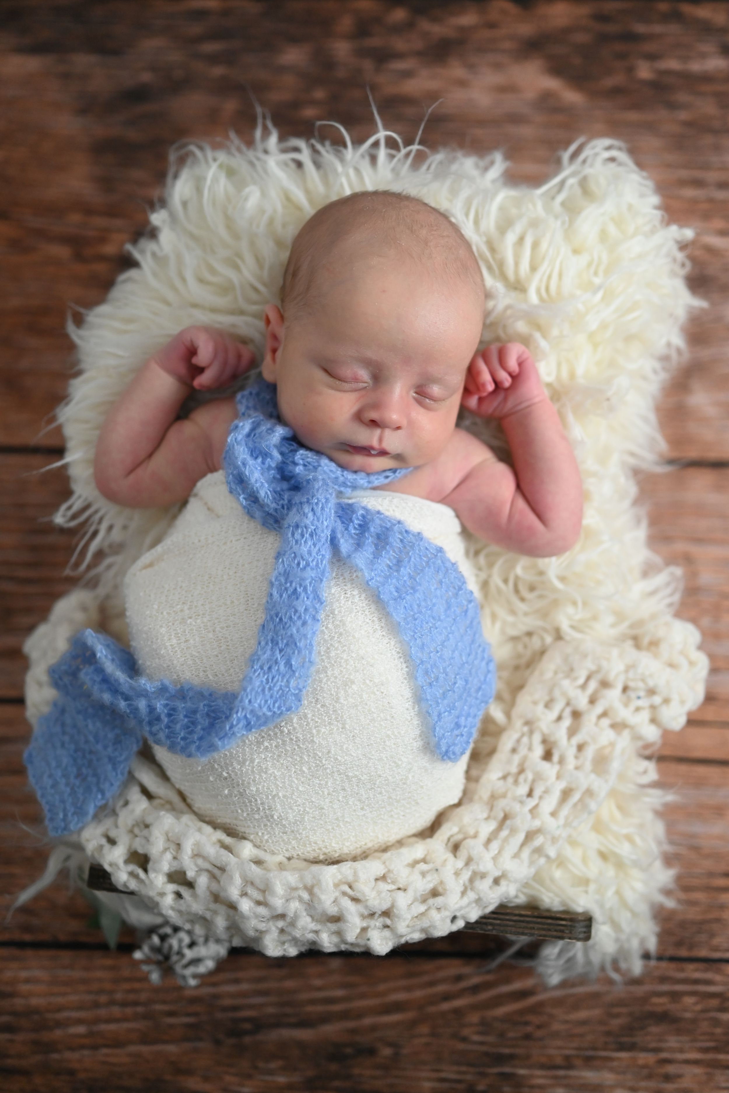 A sleeping newborn swaddled in white and blue lies peacefully on a cream-colored fluffy rug against wooden floorboards.