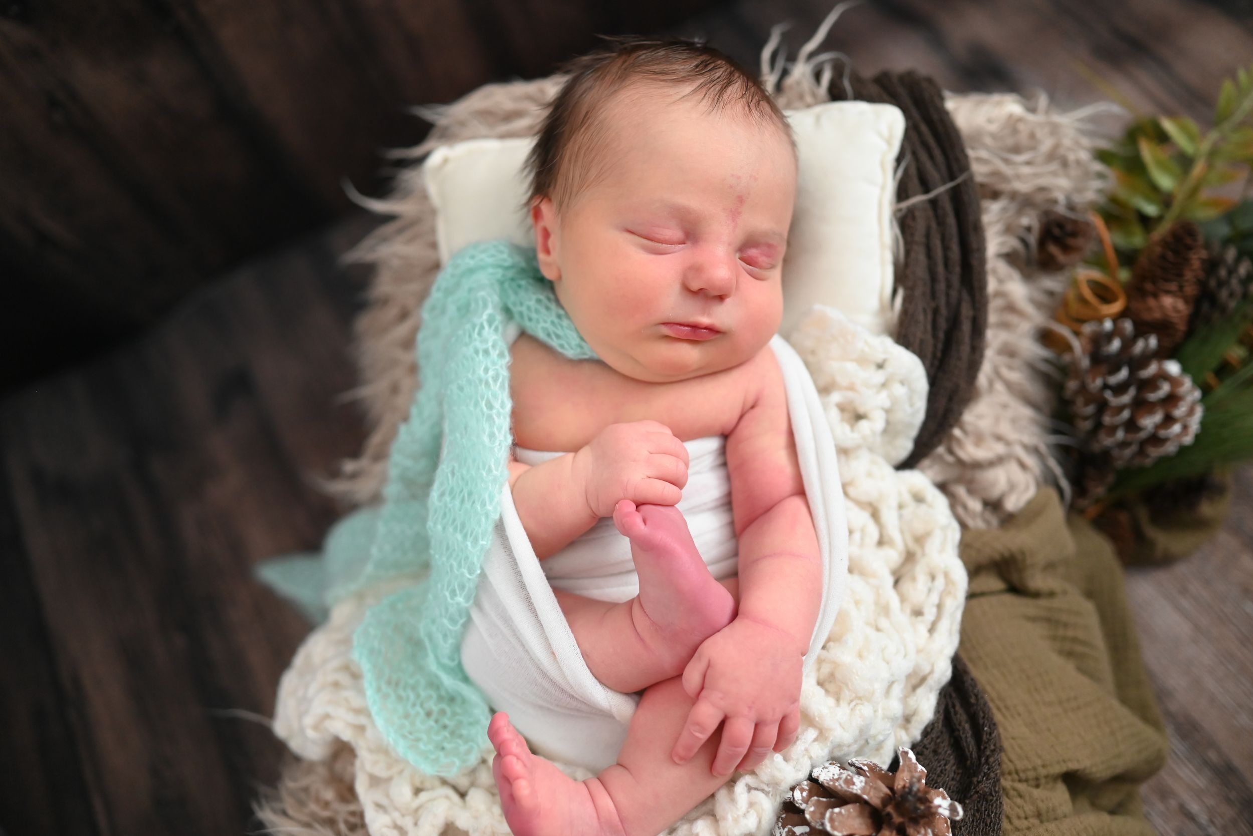 Newborn baby in soft mint green wrap nestled on white blanket with rustic woodland decor.