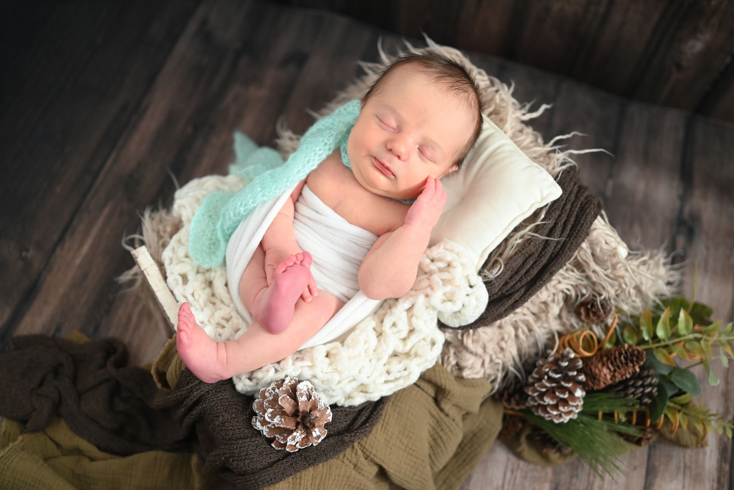 Peaceful newborn sleeping in mint green wrap on white blanket with pinecone decorations.