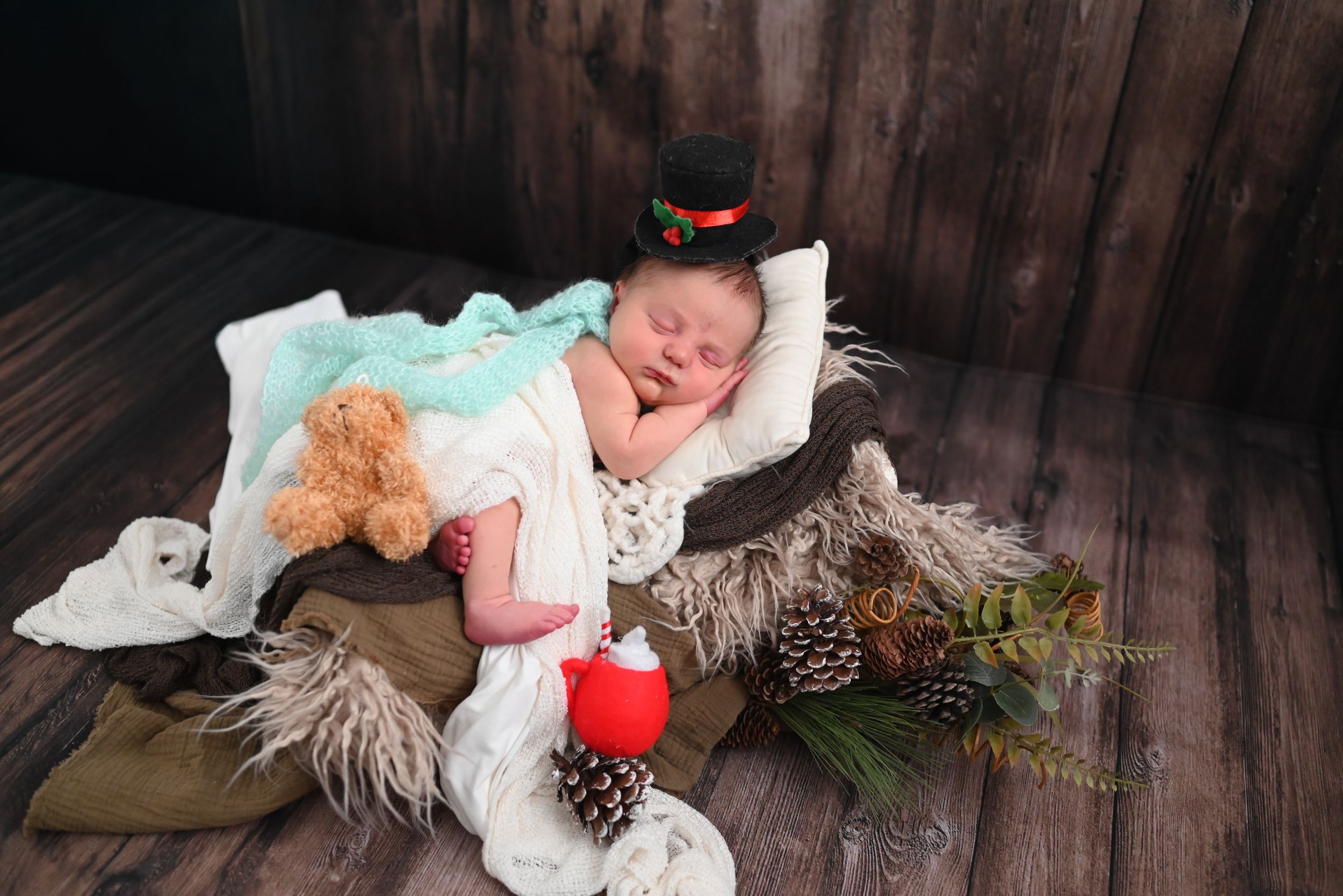 A sleeping infant lies on a rustic wooden backdrop surrounded by pinecones and holiday decorations.