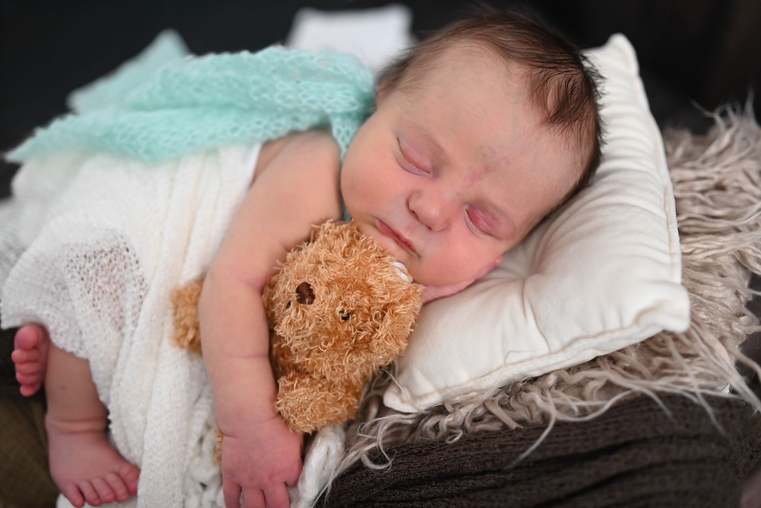 Sleeping baby in aqua blanket cuddling brown teddy bear on white pillow.