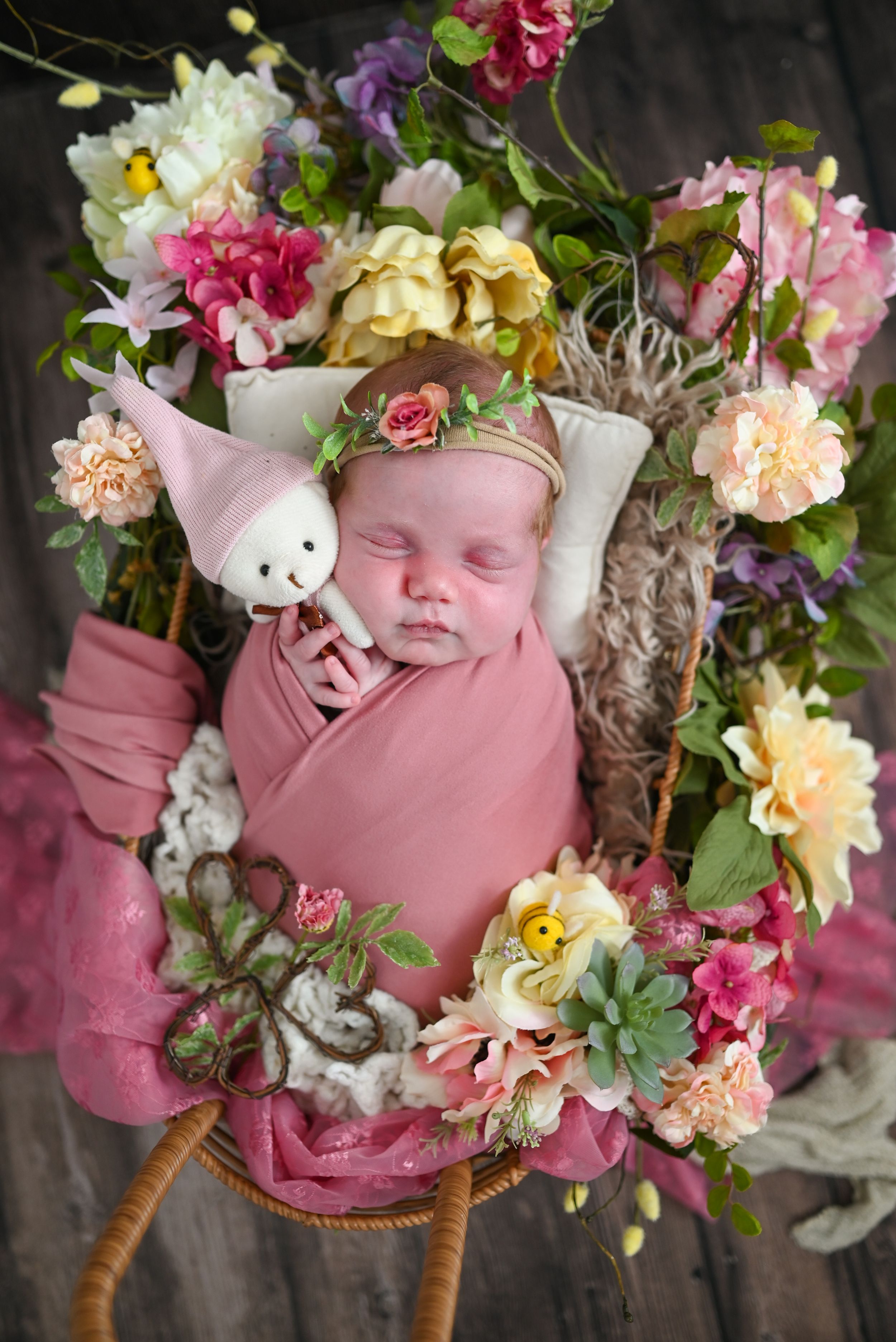 Newborn baby swaddled in pink wrap, sleeping in floral basket with colorful flowers and tiny bunny toy.