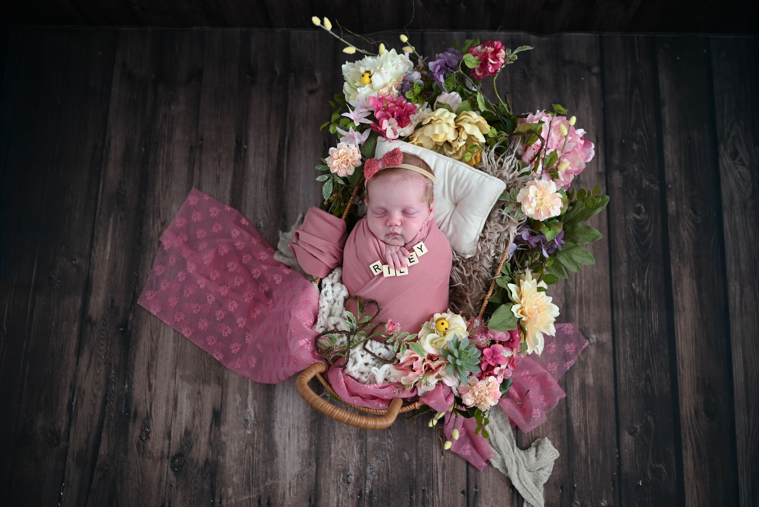 Newborn baby wrapped in pink swaddle surrounded by colorful flowers in a rustic wooden basket.