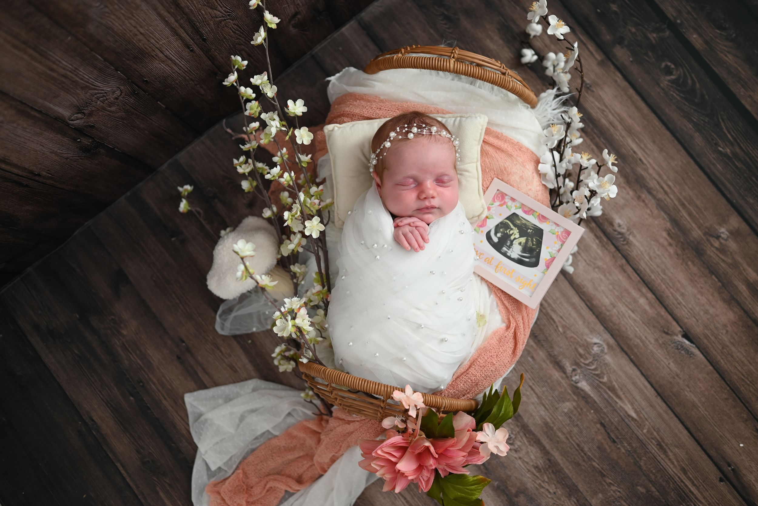 Newborn baby girl wrapped in white lace sleeping in a basket with flowers and ultrasound photo.