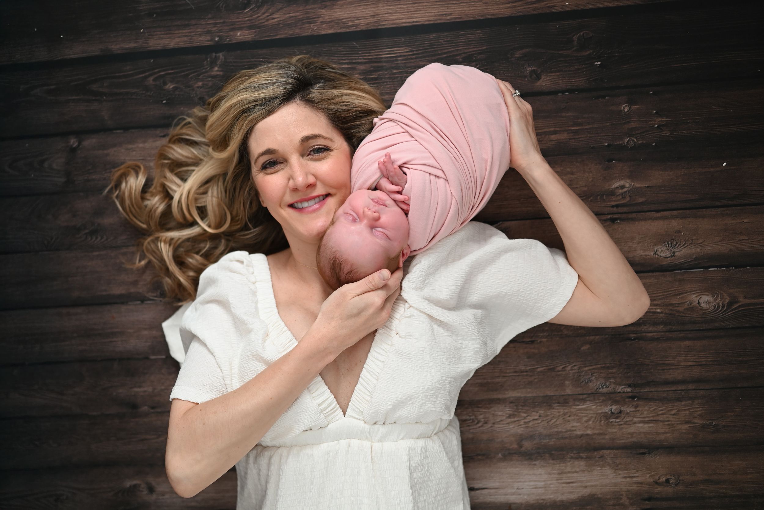 A smiling woman in a white dress lying on wood floor, cradling a newborn baby wrapped in a pink turban.