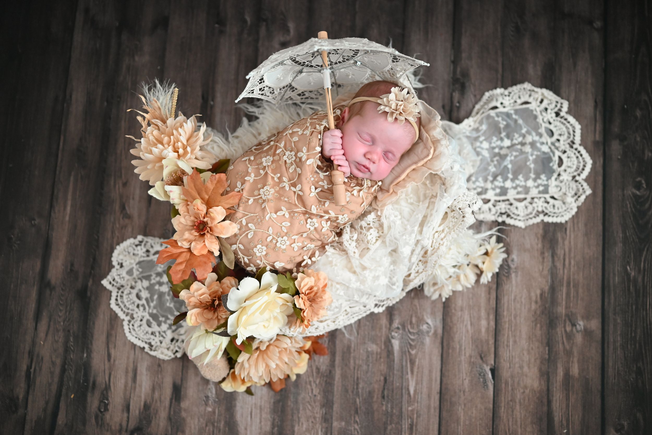 Newborn baby sleeping in lace wrap with floral arrangement and decorative umbrella on rustic wood background.