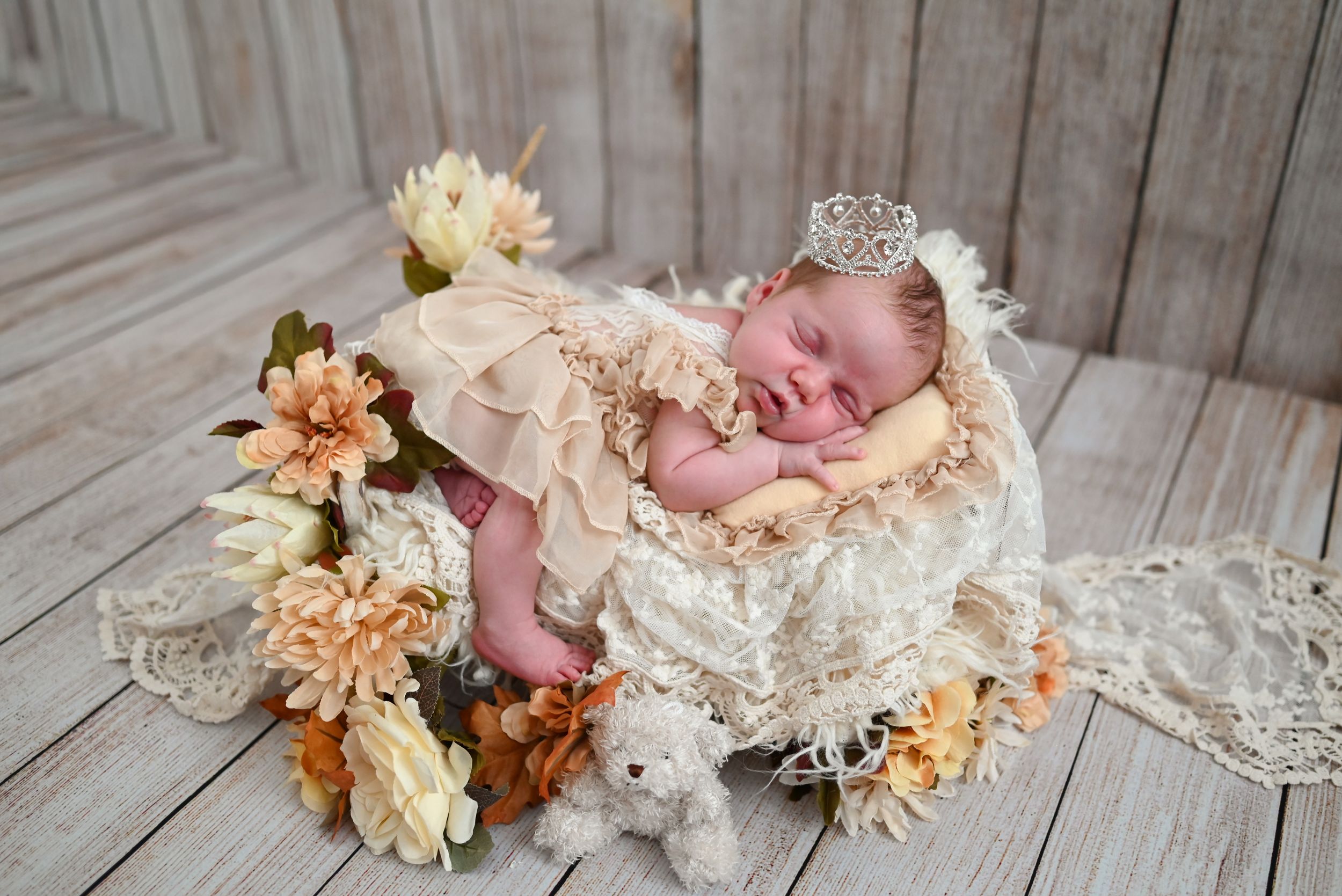 Sleeping newborn baby wearing a tiny crown in a basket surrounded by flowers and a teddy bear.