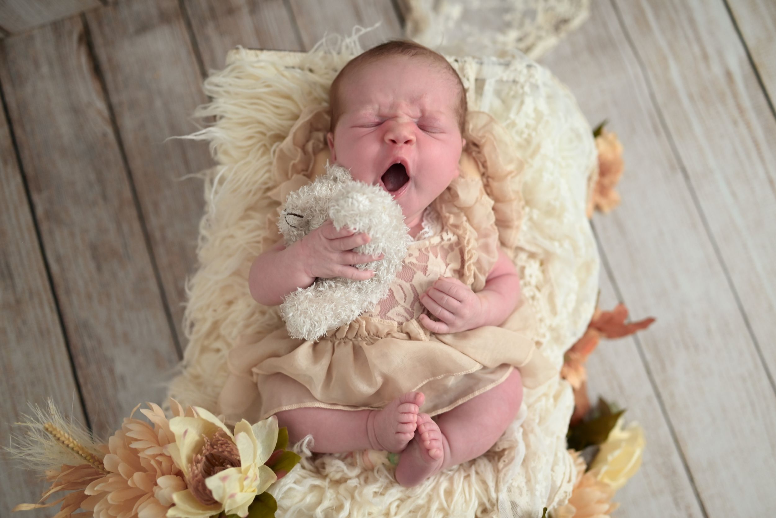 Newborn baby yawning while holding a white stuffed animal, nestled in a fluffy cream basket with flowers.