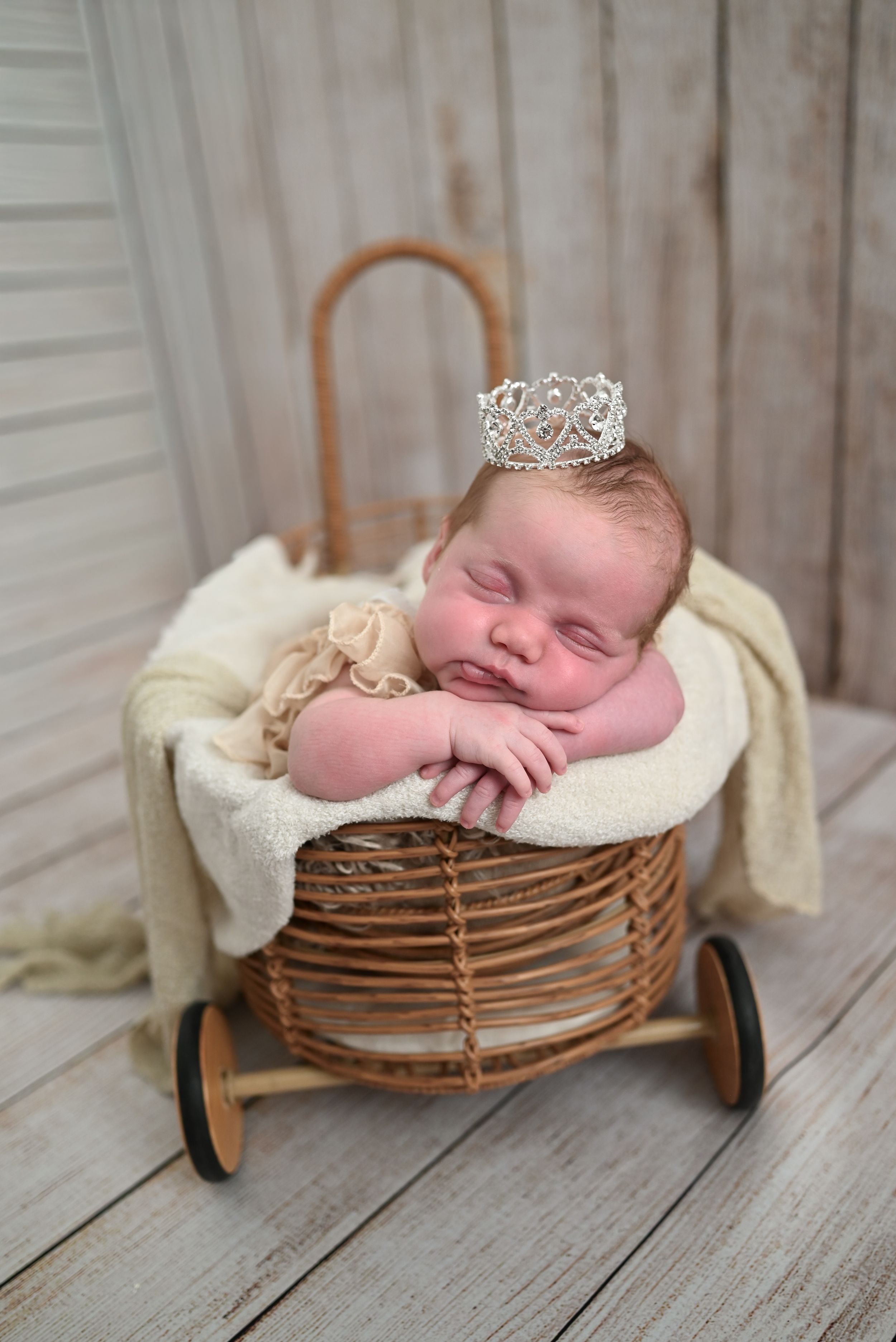 Sleeping newborn baby wearing a silver crown nestled in a wicker basket on wheels with cream blanket.