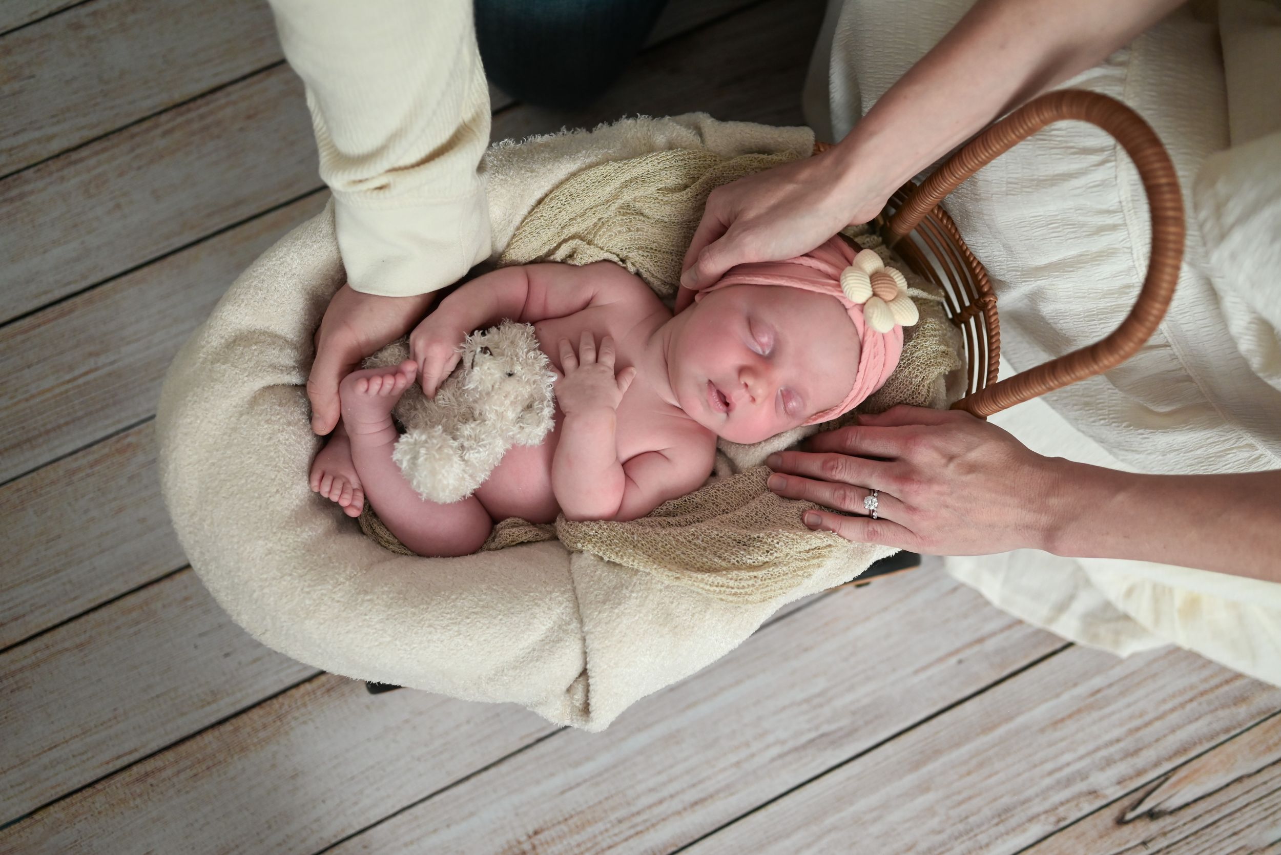 Parents gently cradling sleeping newborn with teddy bear in woven basket on wooden floor.