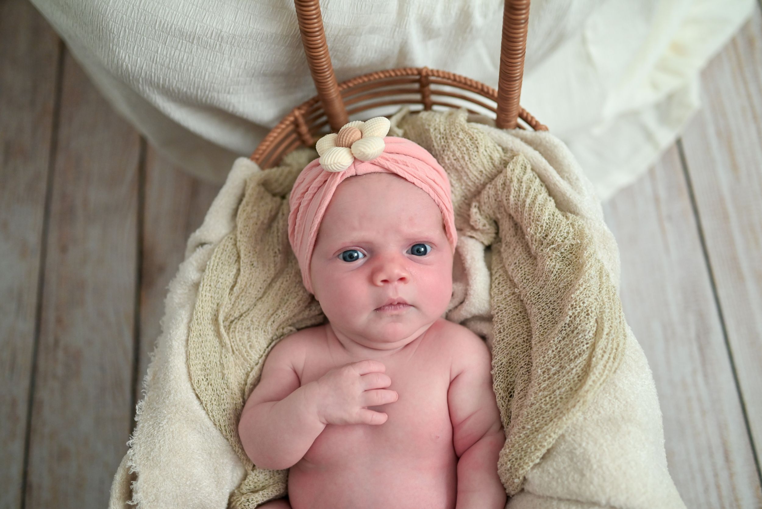 Newborn baby in pink headband lying in wicker basket with cream blanket, wide-eyed and alert.