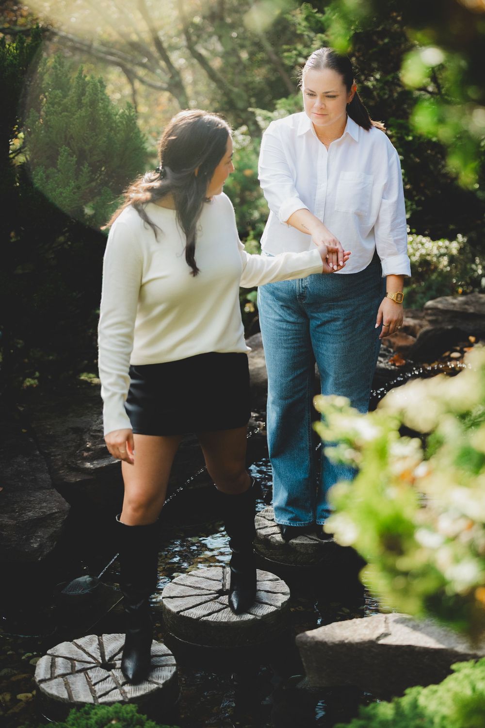 Two people walking on stepping stones in a lush garden path wearing casual outfits in natural lighting.