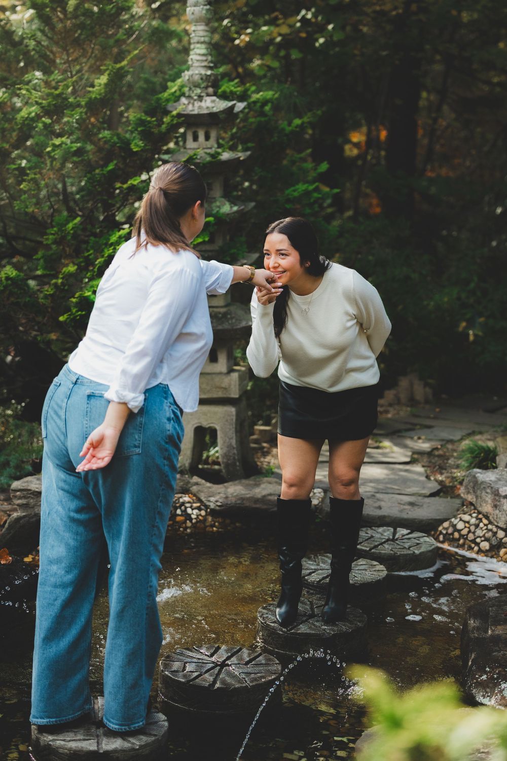 Two people share a playful moment while exploring a tranquil garden stream with decorative stone features.