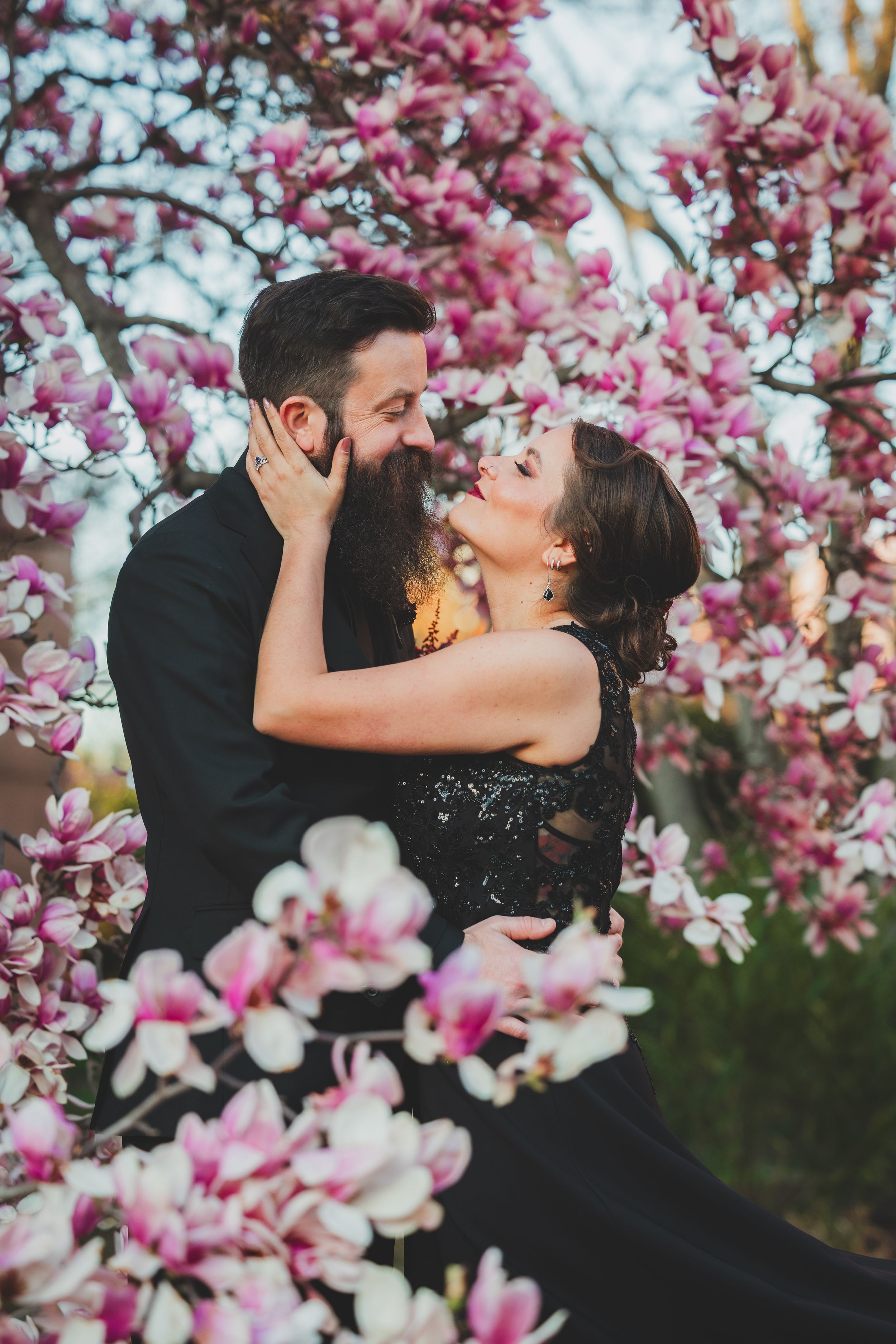 A romantic couple shares an intimate moment surrounded by blooming pink magnolia blossoms in springtime.