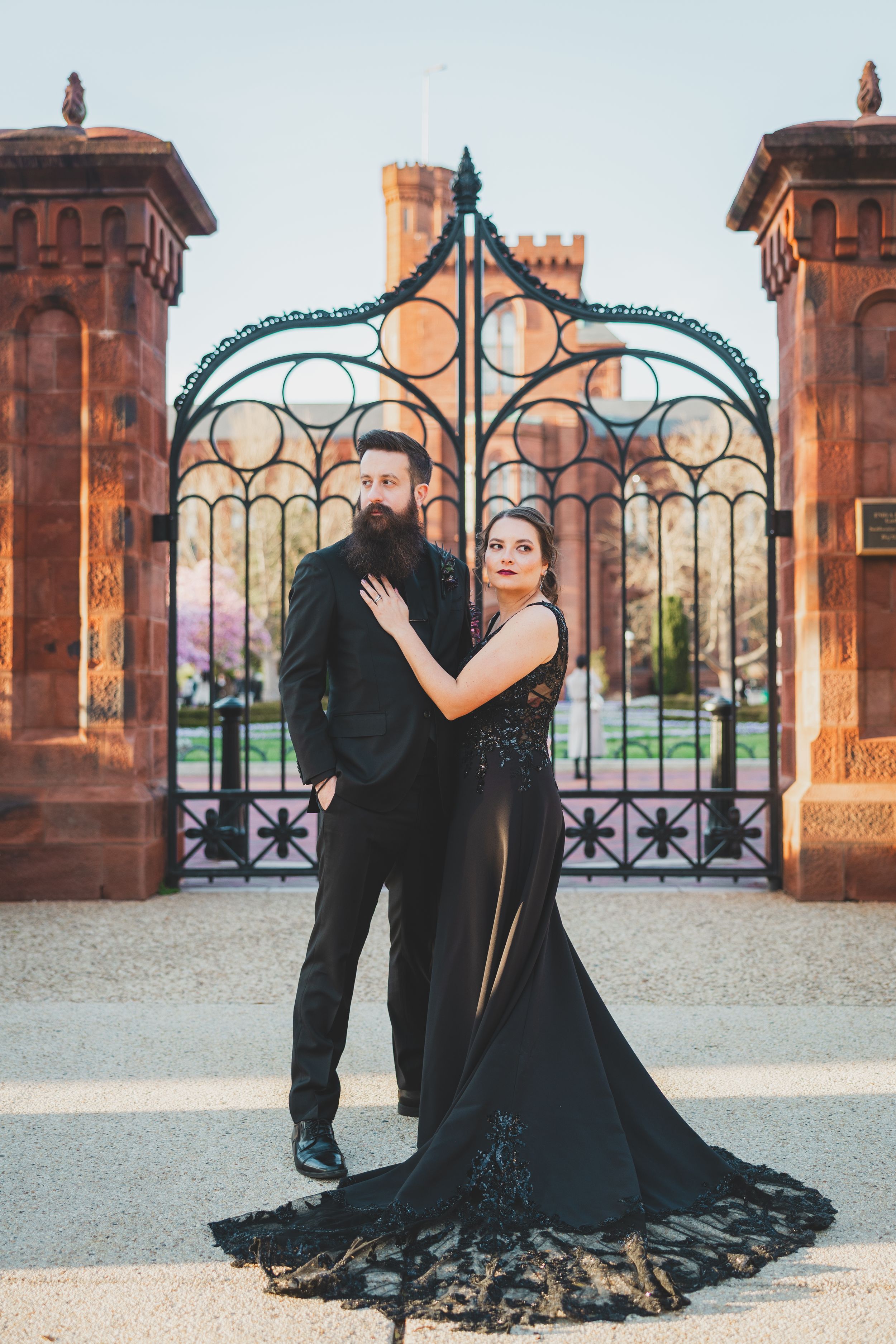 Elegant couple in black formal attire pose in front of ornate iron gates and stone pillars of the Enid Haupt