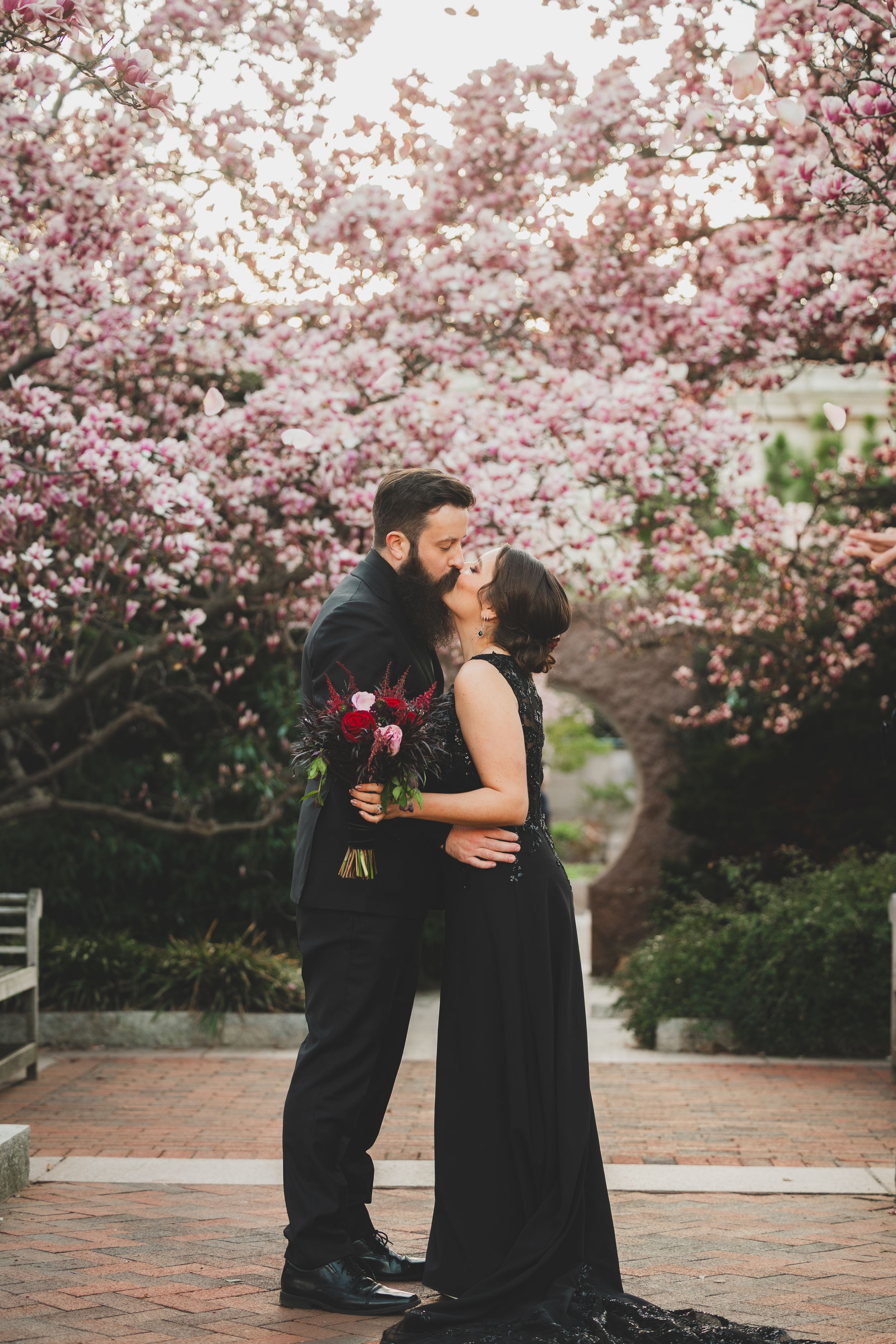 A romantic couple shares a tender moment beneath blooming pink cherry blossoms in a garden setting.