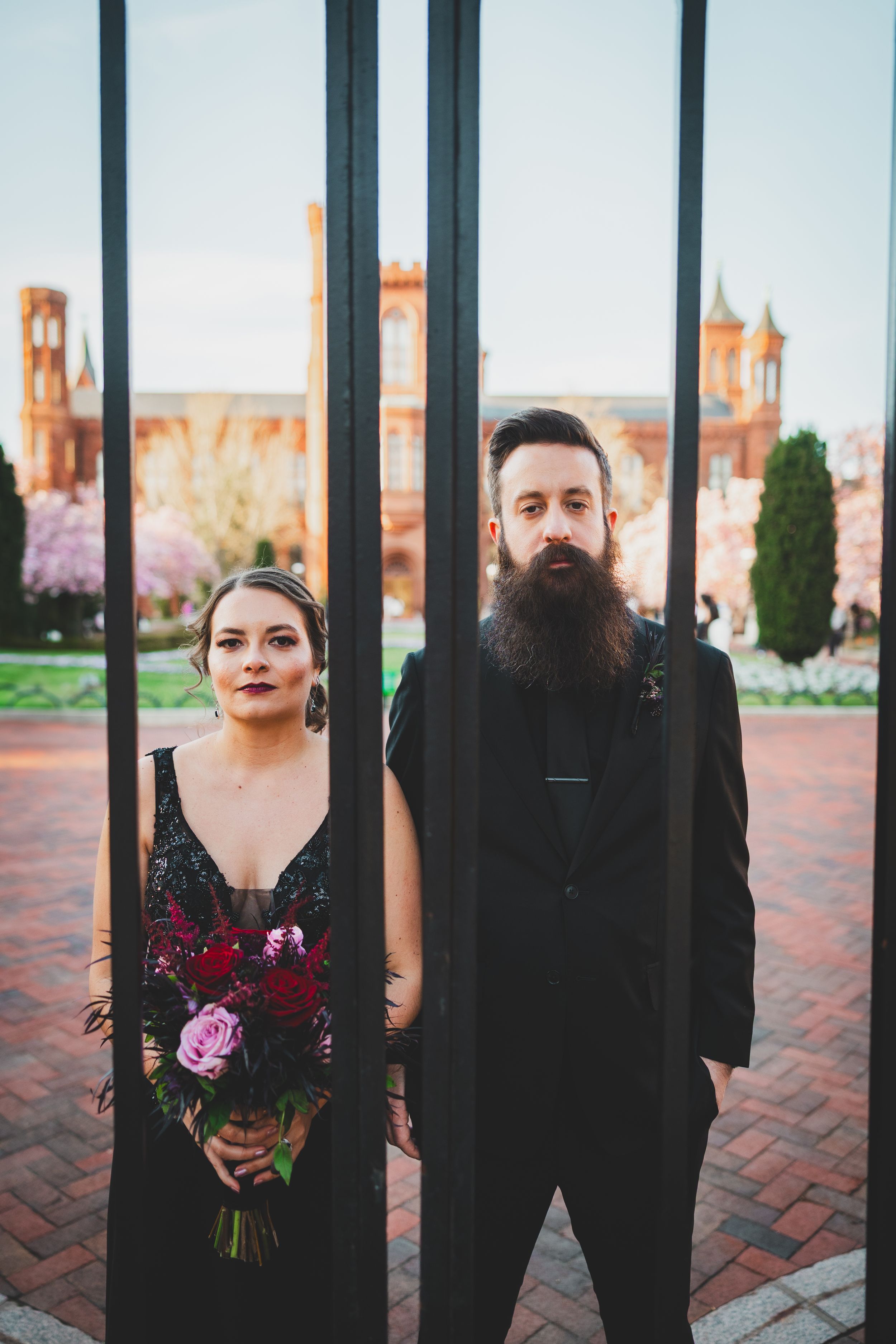 A couple in black formal attire stands behind iron bars with gothic architecture visible in the background.
