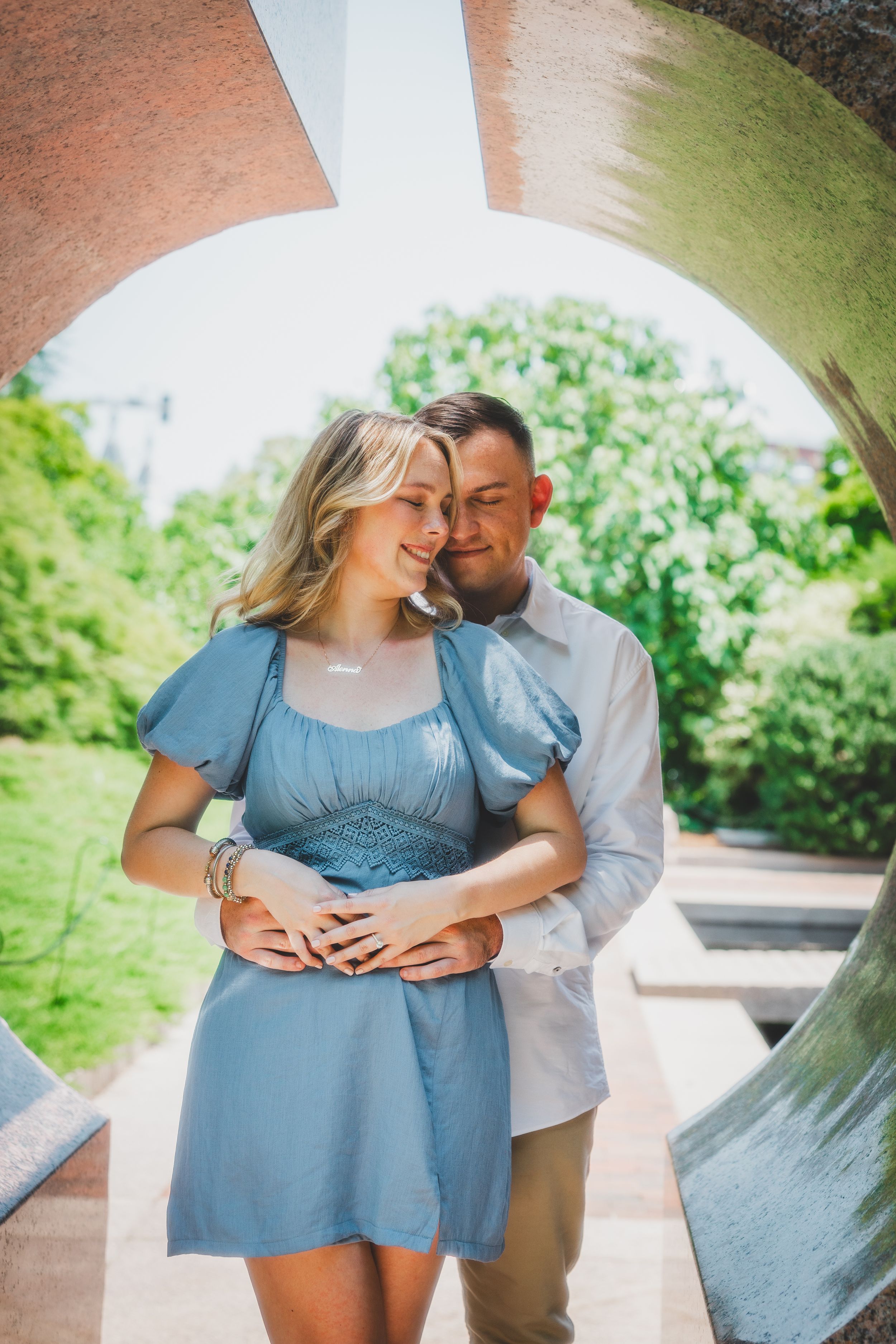 Couple in coordinated blue and white outfits embrace lovingly outdoors in a lush garden setting during a proposal photoshoot at Enid Haupt