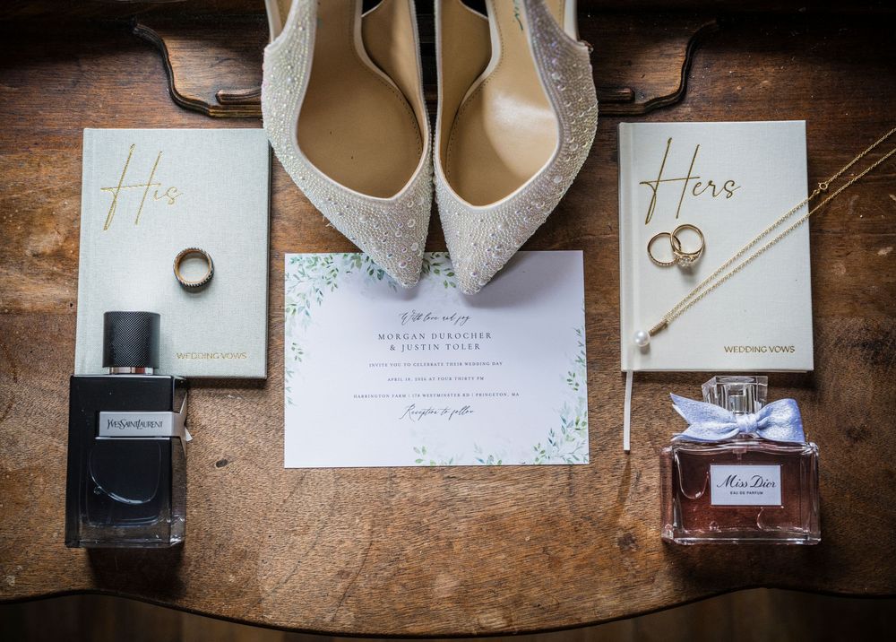 Elegant wedding flat lay featuring glittery heels, His and Hers books, gold jewelry, perfume bottles, and a floral invitation on wood.