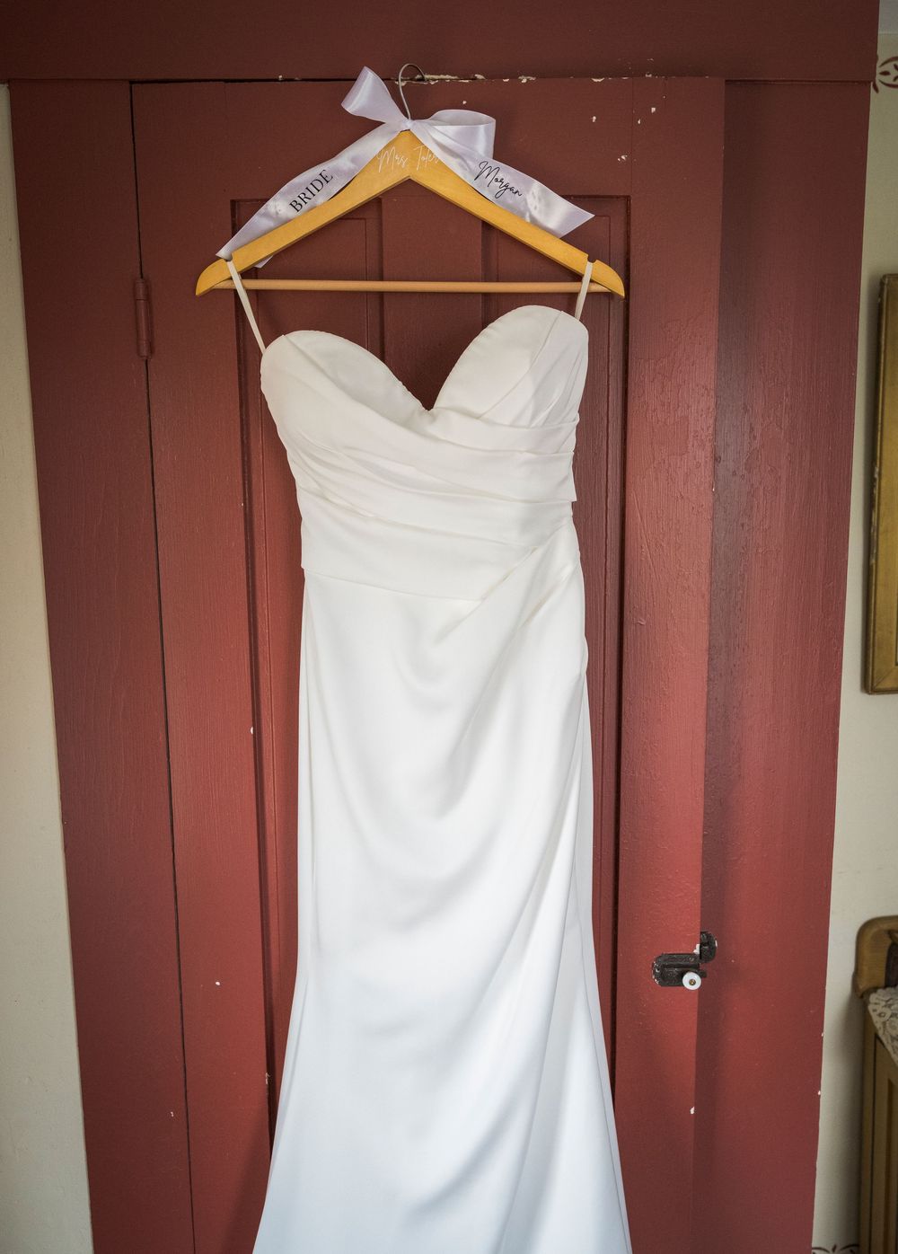White strapless mermaid wedding dress hanging on a personalized wooden hanger against a red door.