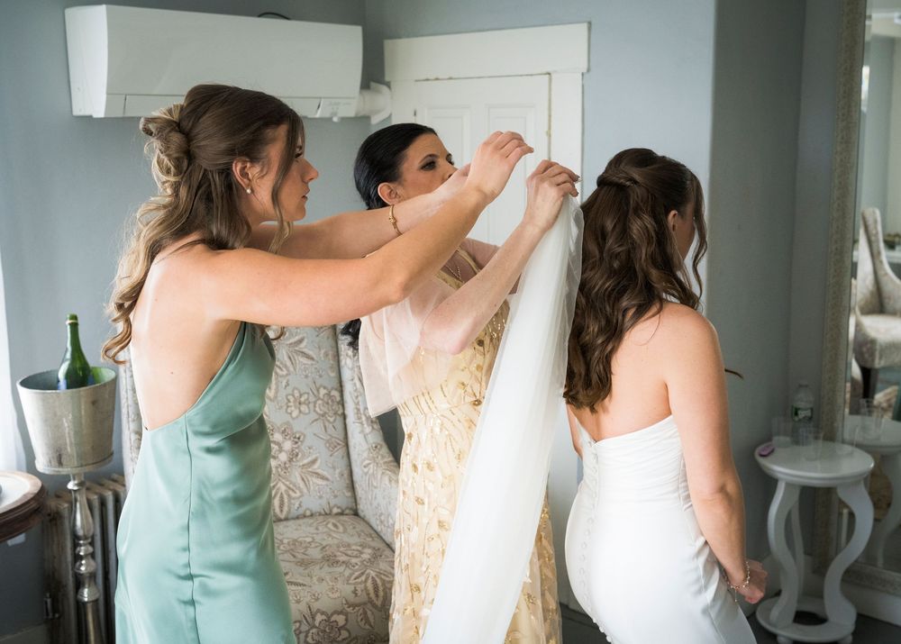 Bridesmaids helping bride into white wedding gown in elegant room with champagne bottle nearby.