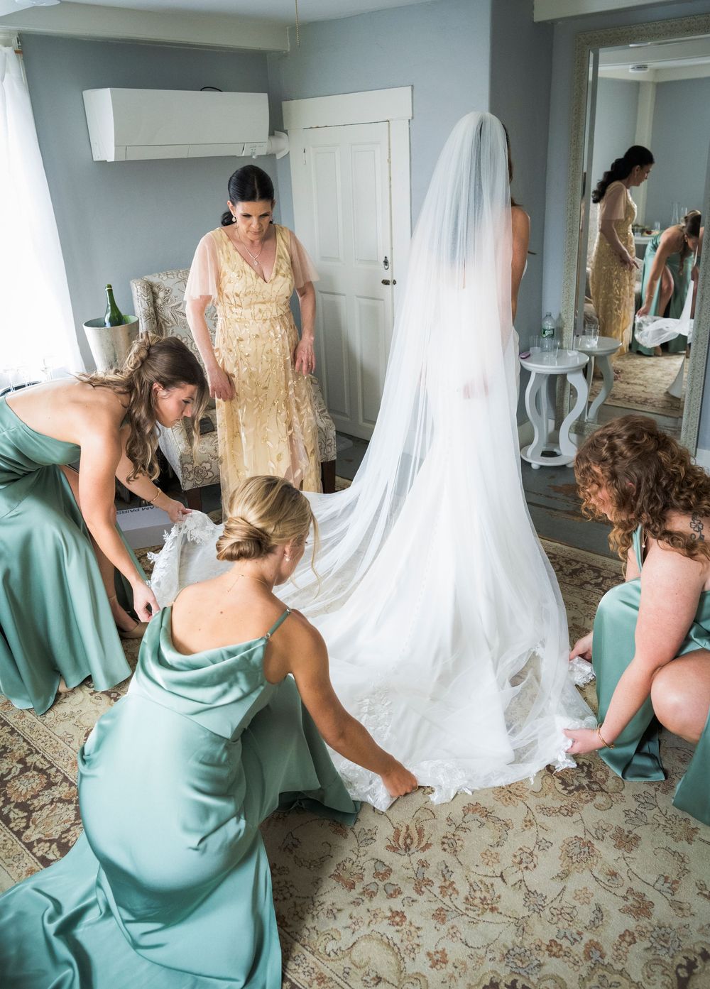 Bridesmaids in sage green gowns help bride into wedding dress while woman in gold dress assists in elegant room.